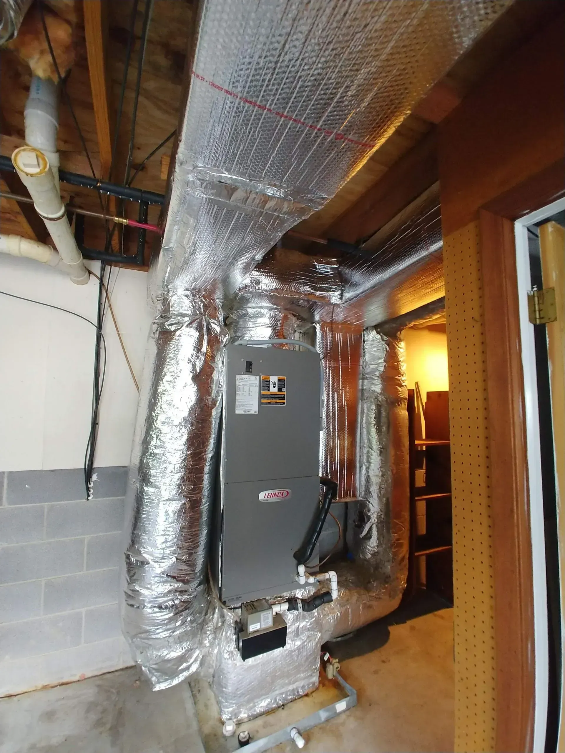 Furnace and ductwork in a basement. Silver insulated ducts are attached to a grey Lennox furnace.