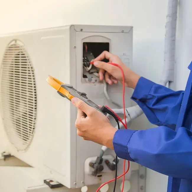 HVAC technician in blue overalls tests an air conditioning unit's wiring with a multimeter.