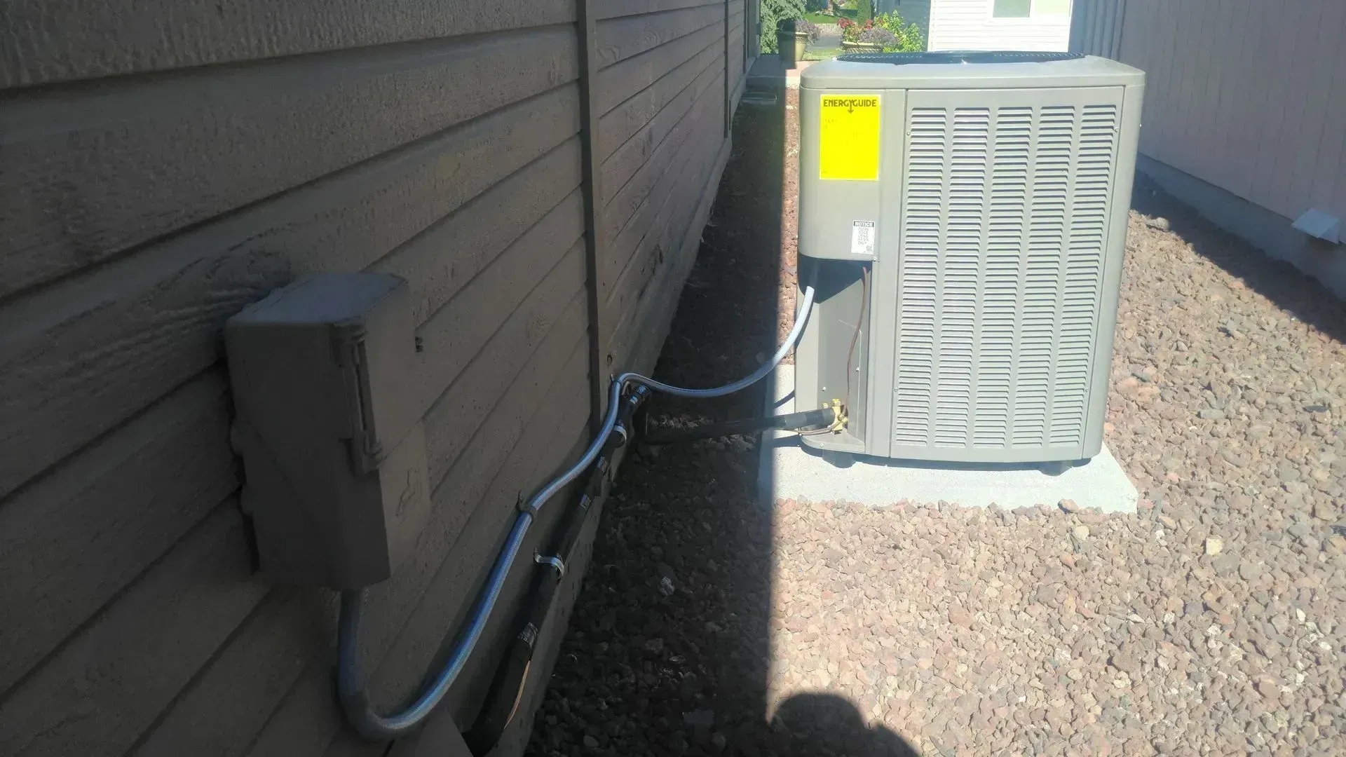 Air conditioner unit next to a house wall with electrical box and gravel ground.