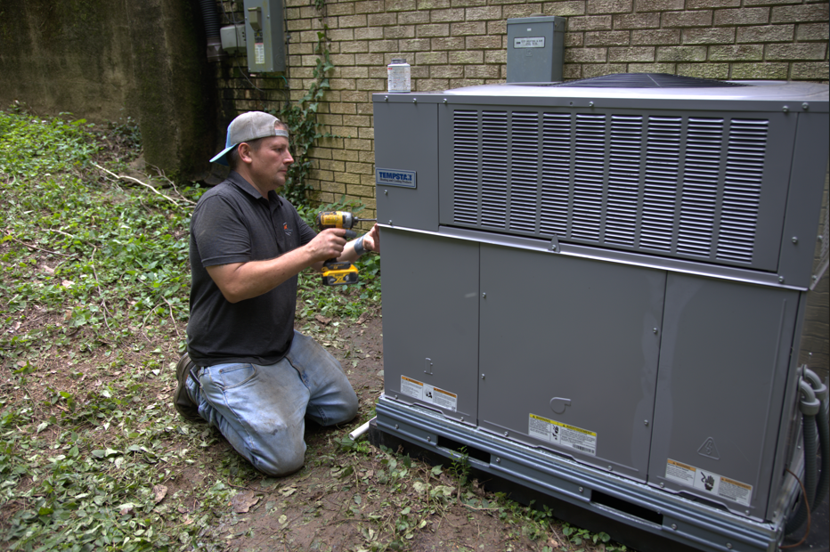 A person in a baseball cap kneels, using a power drill on an outdoor HVAC unit, near a brick wall.