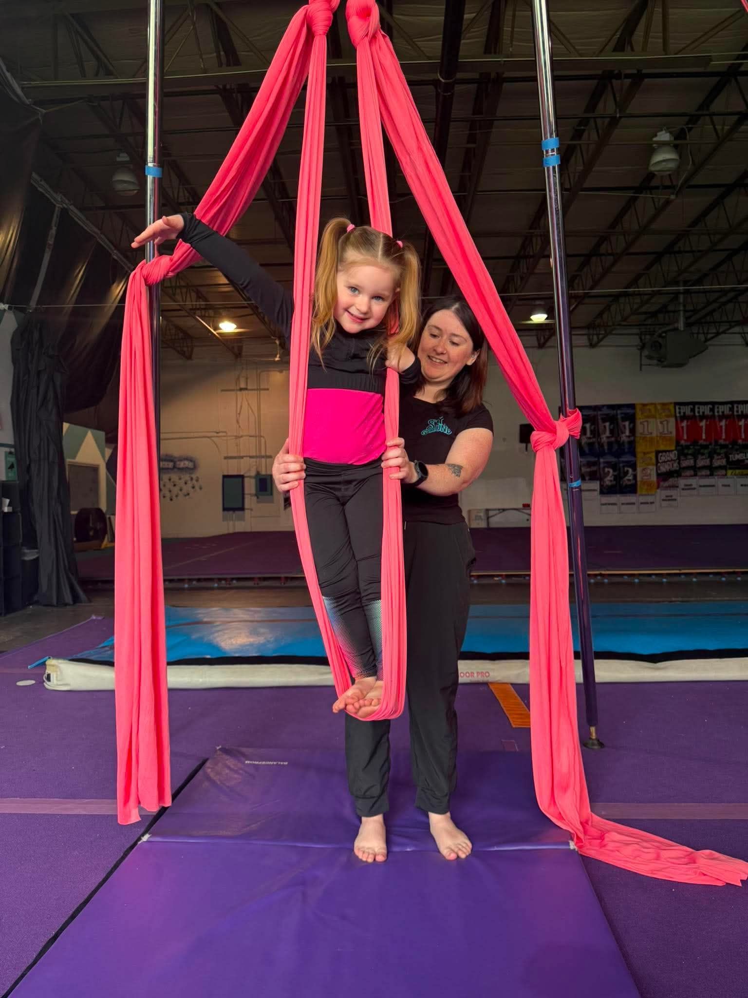 A child in a green t-shirt performs aerial yoga, suspended in a bright red fabric swing inside a gym.