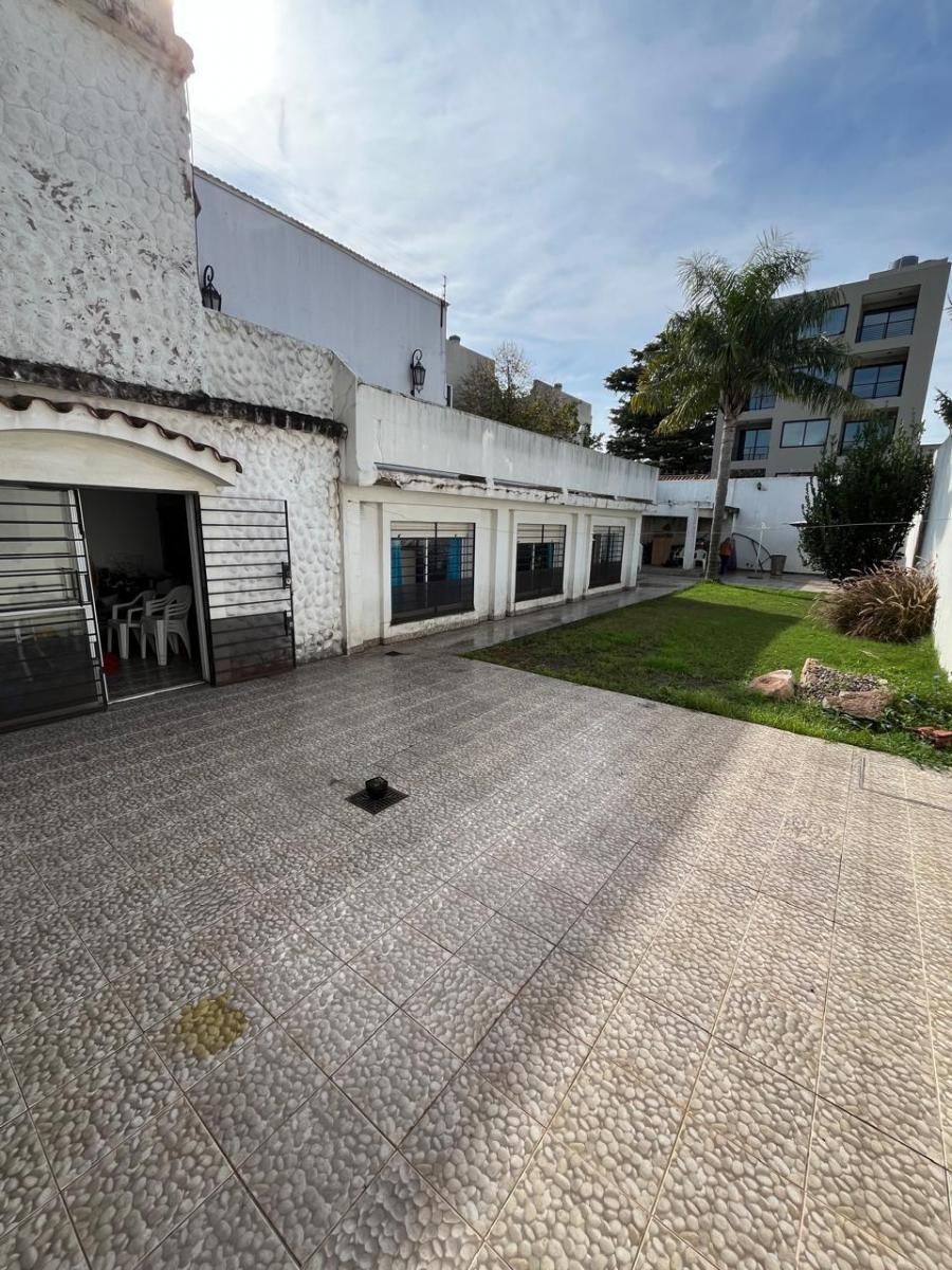 Patio vacío con pavimento de baldosas, edificio blanco y una pequeña zona de césped bajo un cielo nublado.