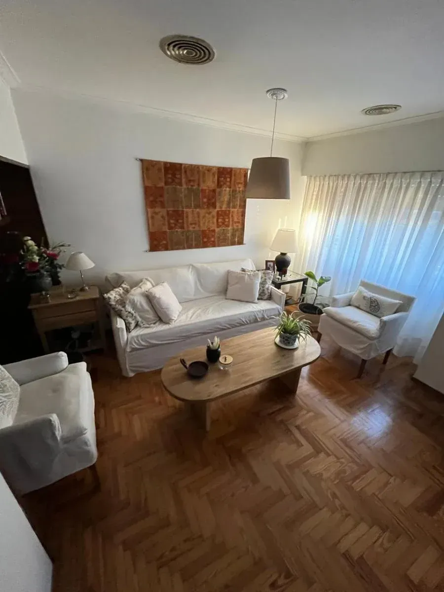 Luminoso salón con sofá blanco, sillones, mesa de madera, suelo estampado y cortinas transparentes.