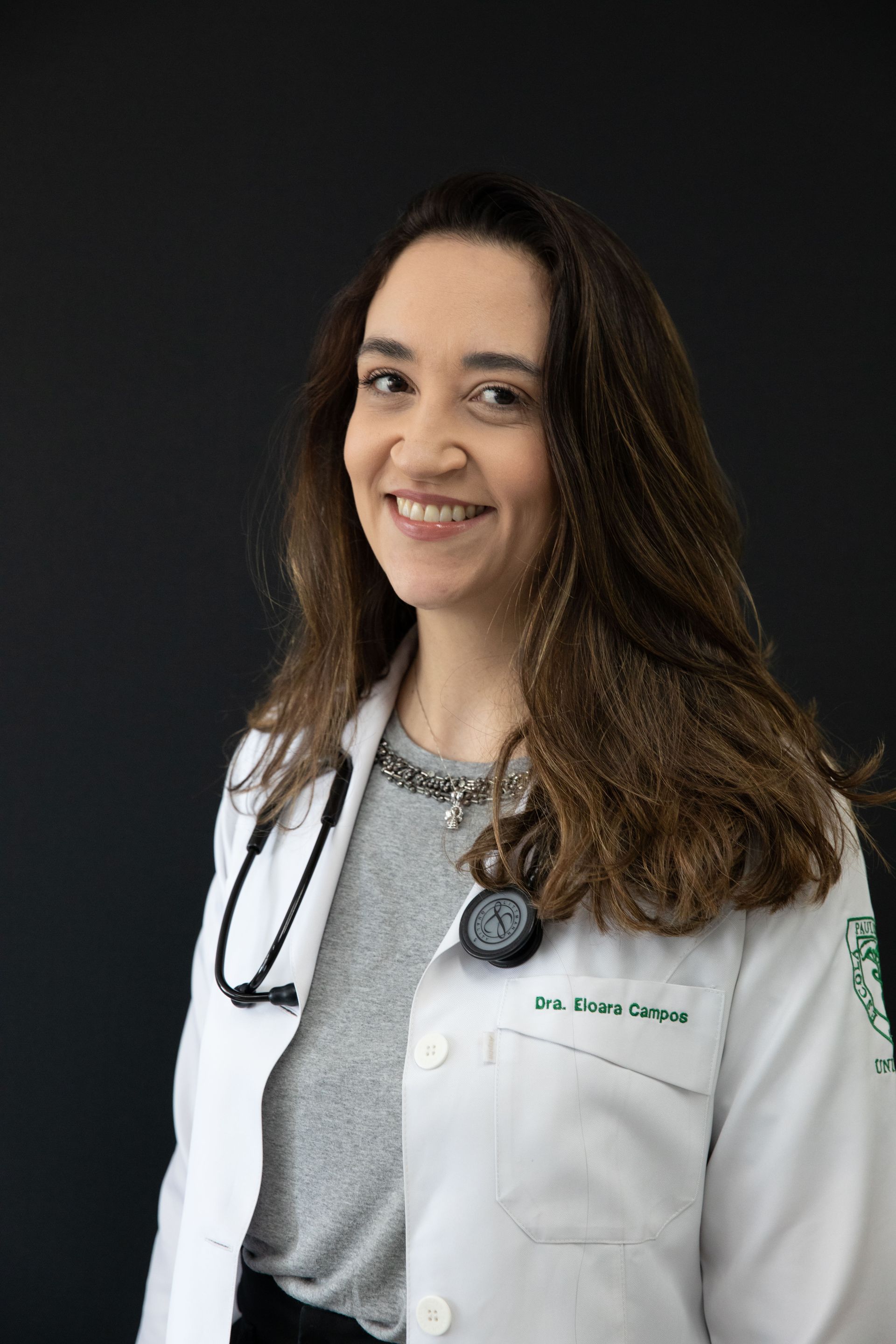 Uma mulher de jaleco com um estetoscópio no pescoço está sorrindo.