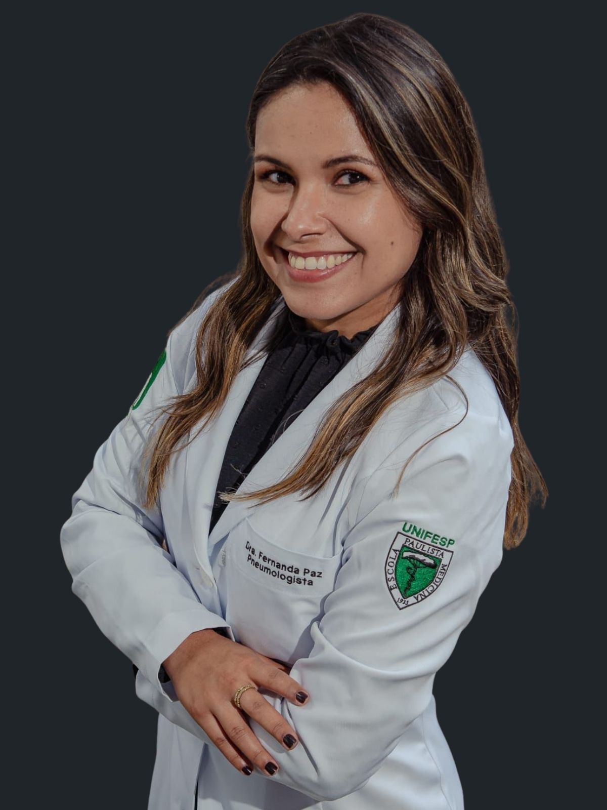 Uma mulher de jaleco branco está sorrindo com os braços cruzados.