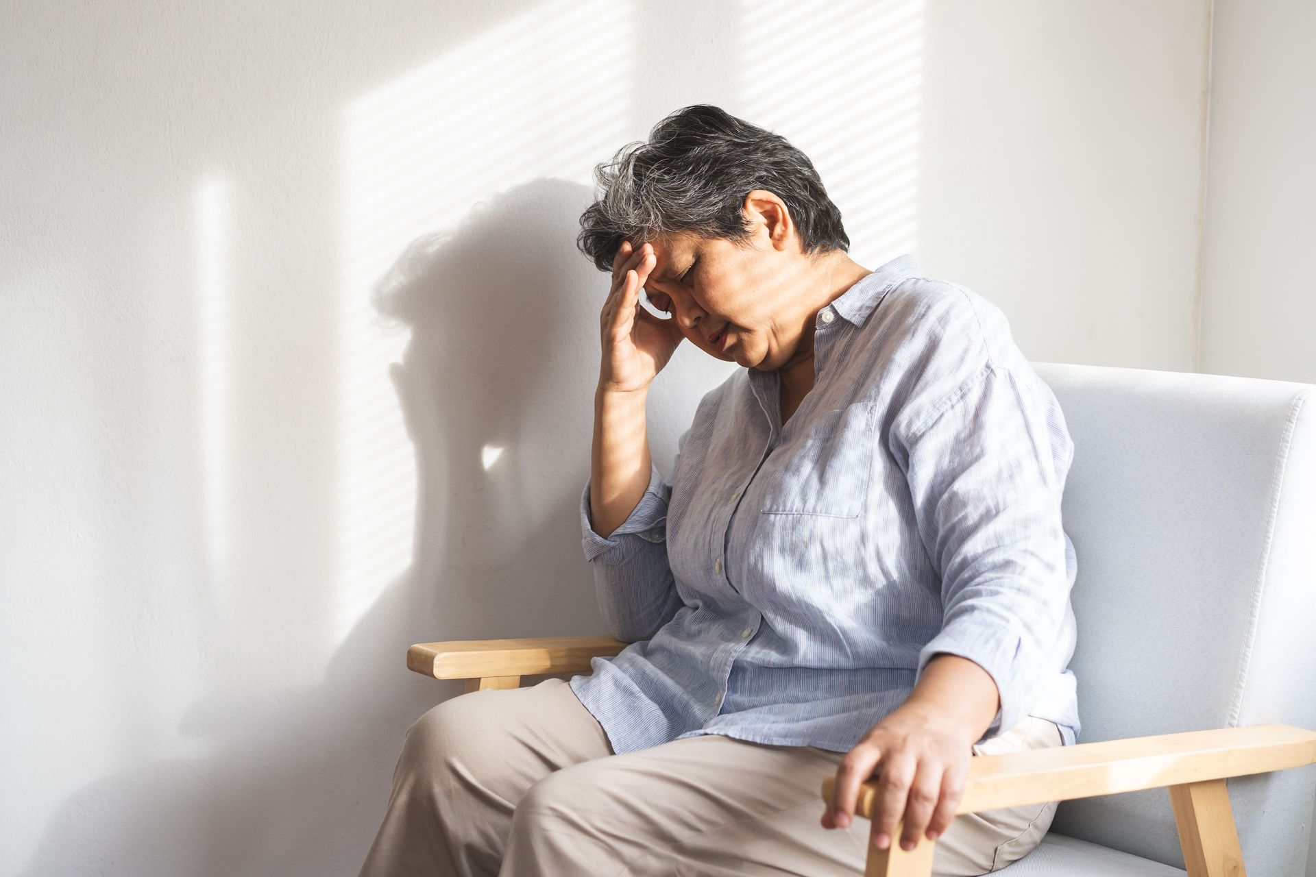 Uma senhora idosa está sentada em uma cadeira com a cabeça entre as mãos.