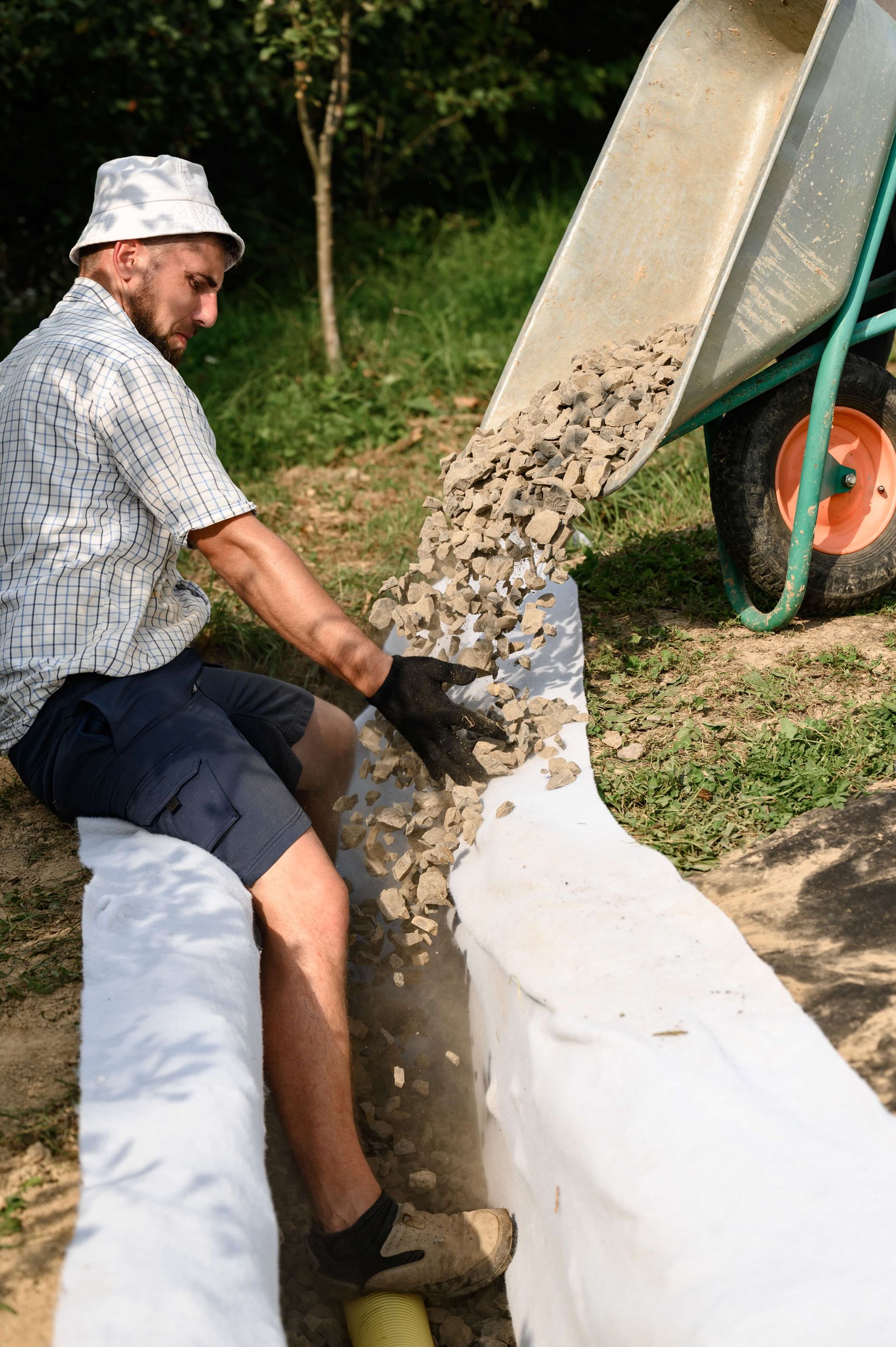 Man emptying gravel from a wheelbarrow into a trench in a garden.