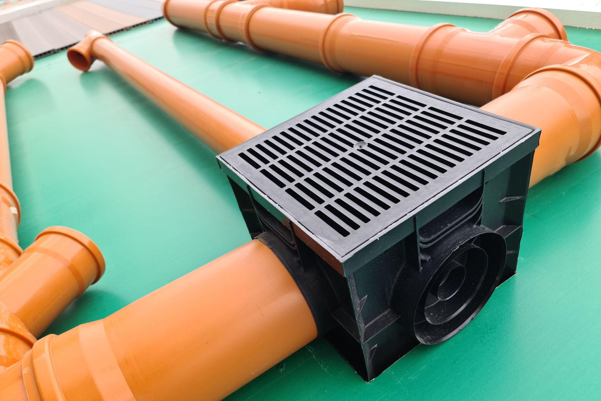 Black drain with grate connected to orange PVC pipes on a green surface.