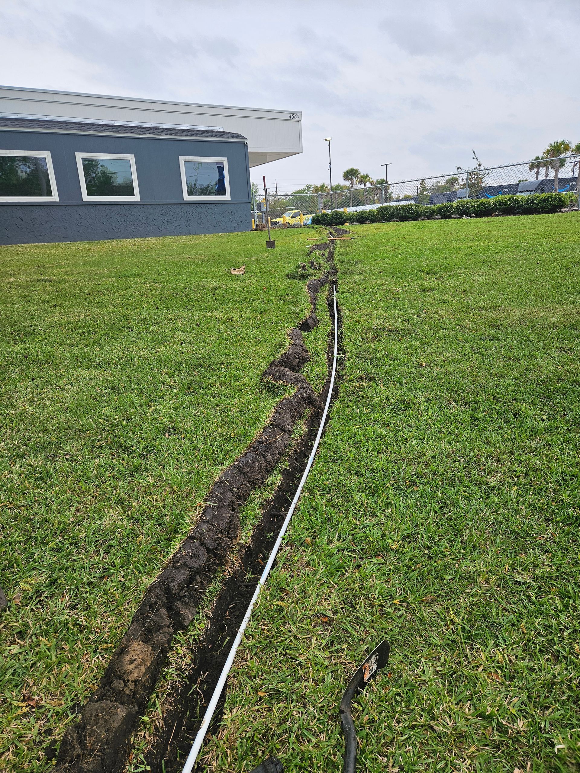 Trench dug in grass, with a black cable running along it, beside a gray building with windows.
