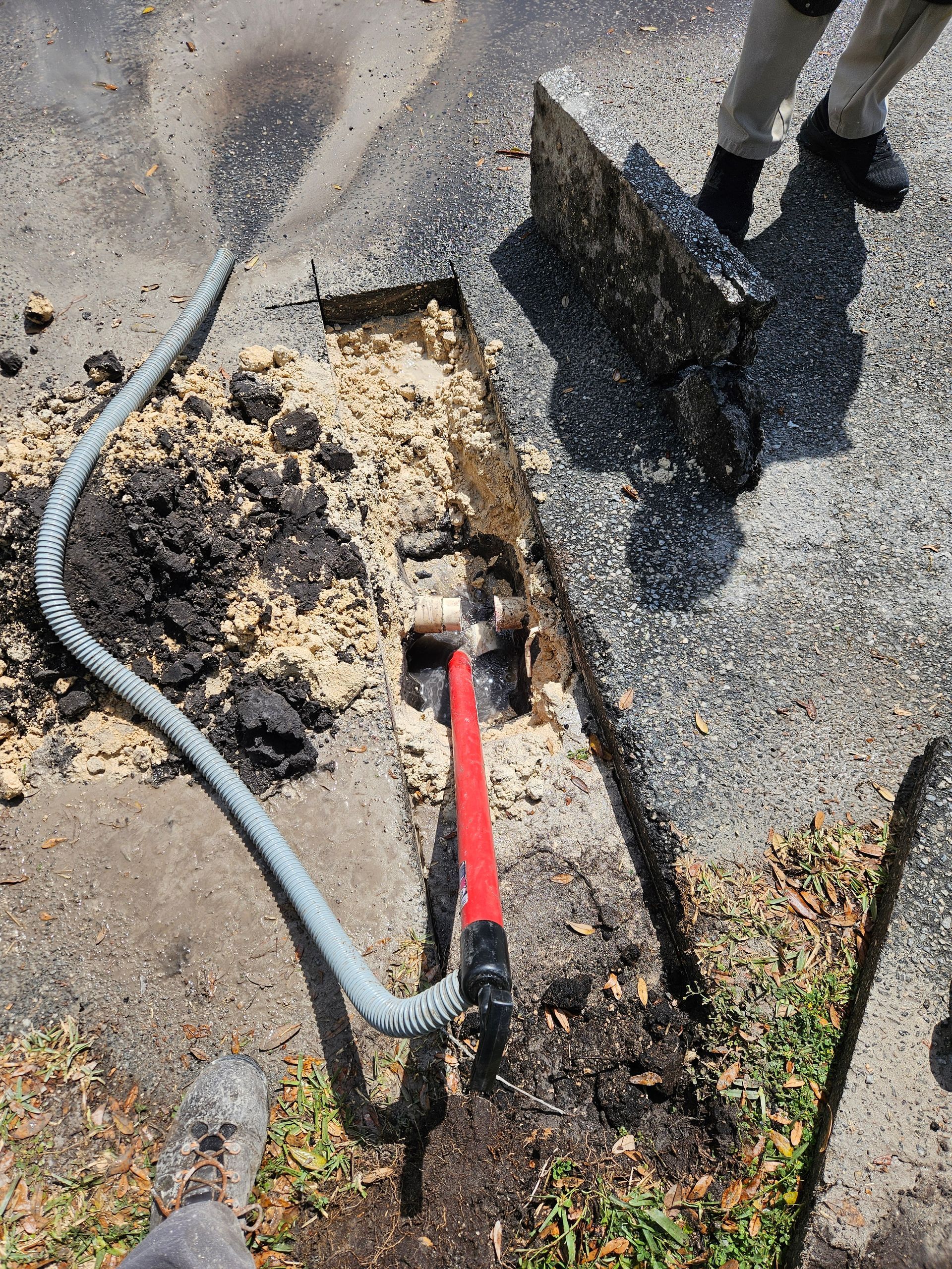 Workers excavating asphalt with a hammer and hose. Asphalt fragments and light-colored dirt are visible.