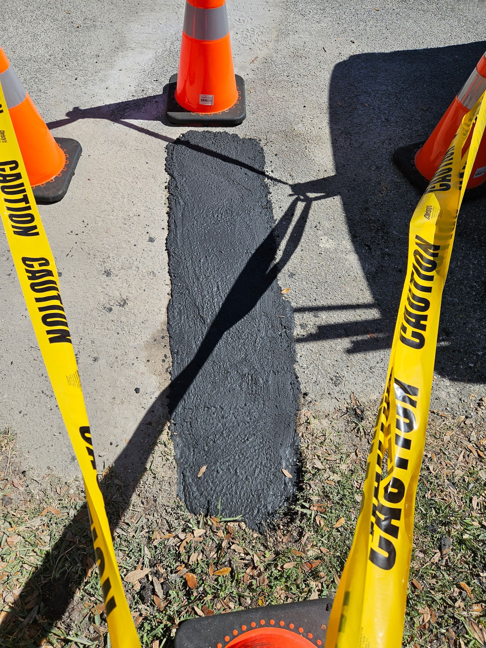Freshly paved asphalt patch bordered by caution tape and traffic cones.