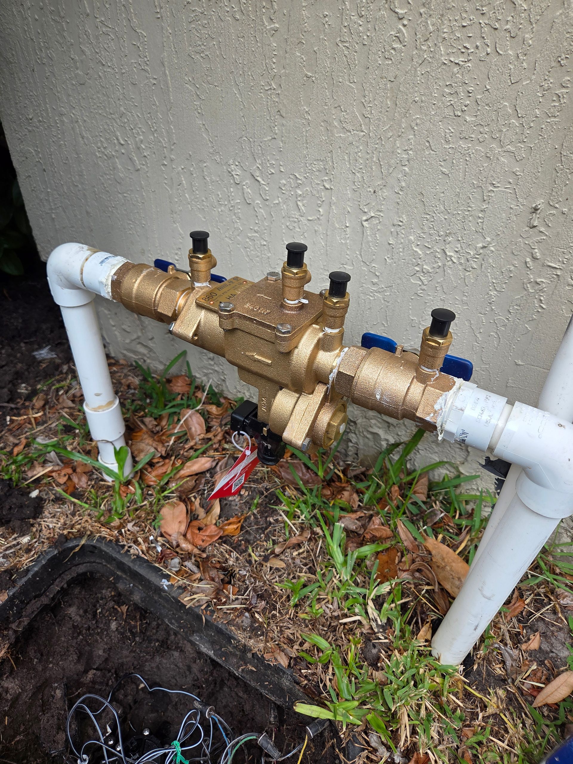 A backflow preventer with brass body and PVC pipes near a wall and grass.