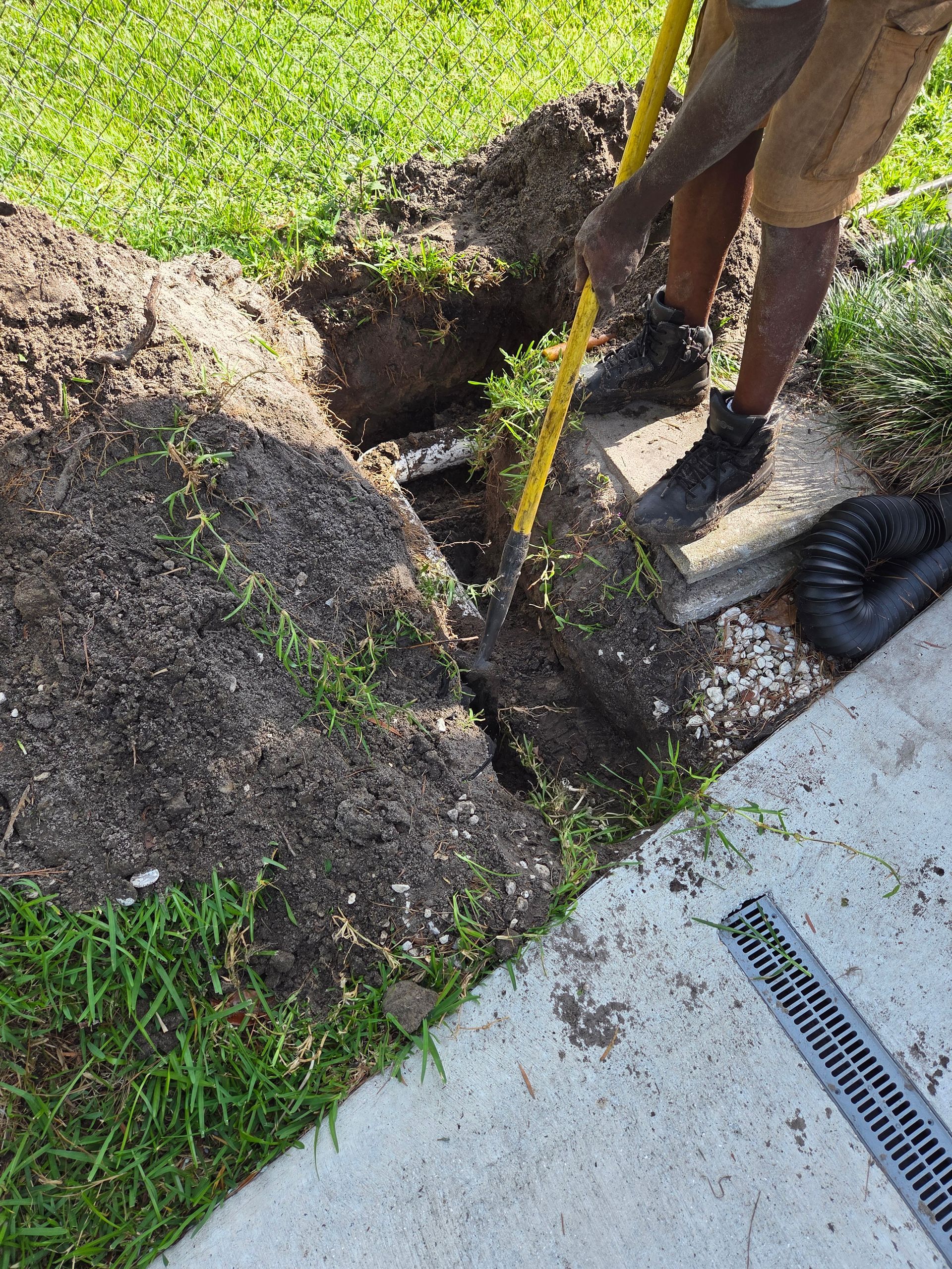 Person digging in a trench with a shovel next to a concrete walkway and drain.