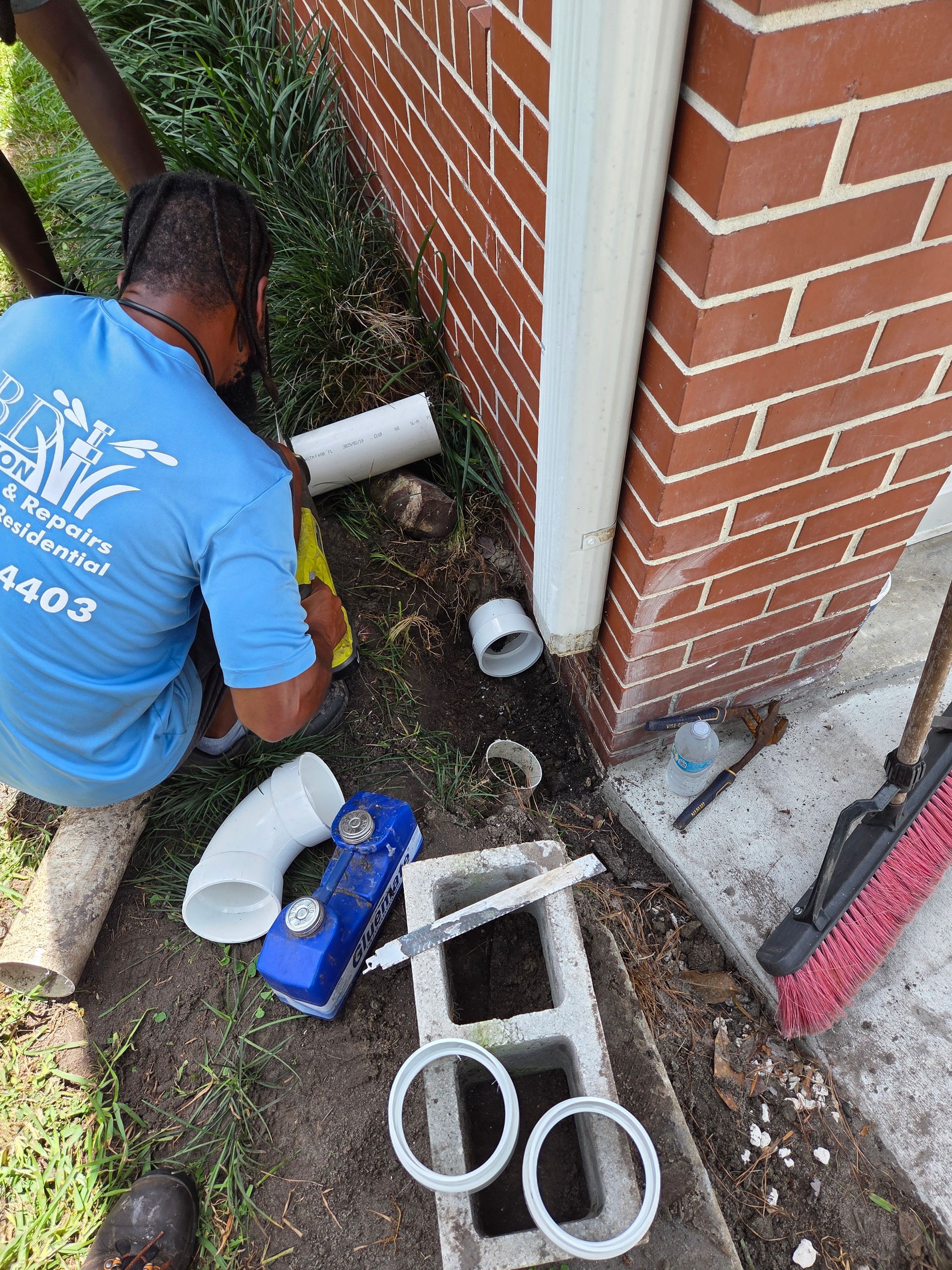 Person installing drainage pipe near a brick building. Parts, tools, and a broom are nearby.