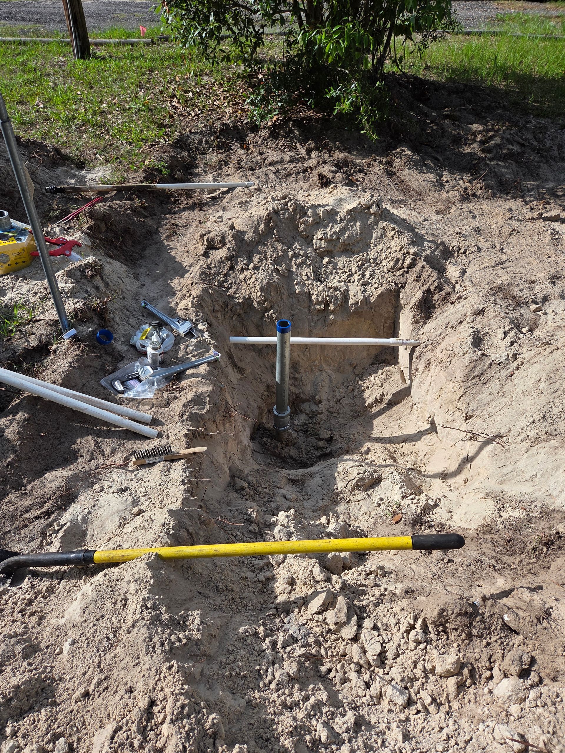 Hole dug in dirt with metal bars, piping, and a yellow shovel. Construction or repair site.