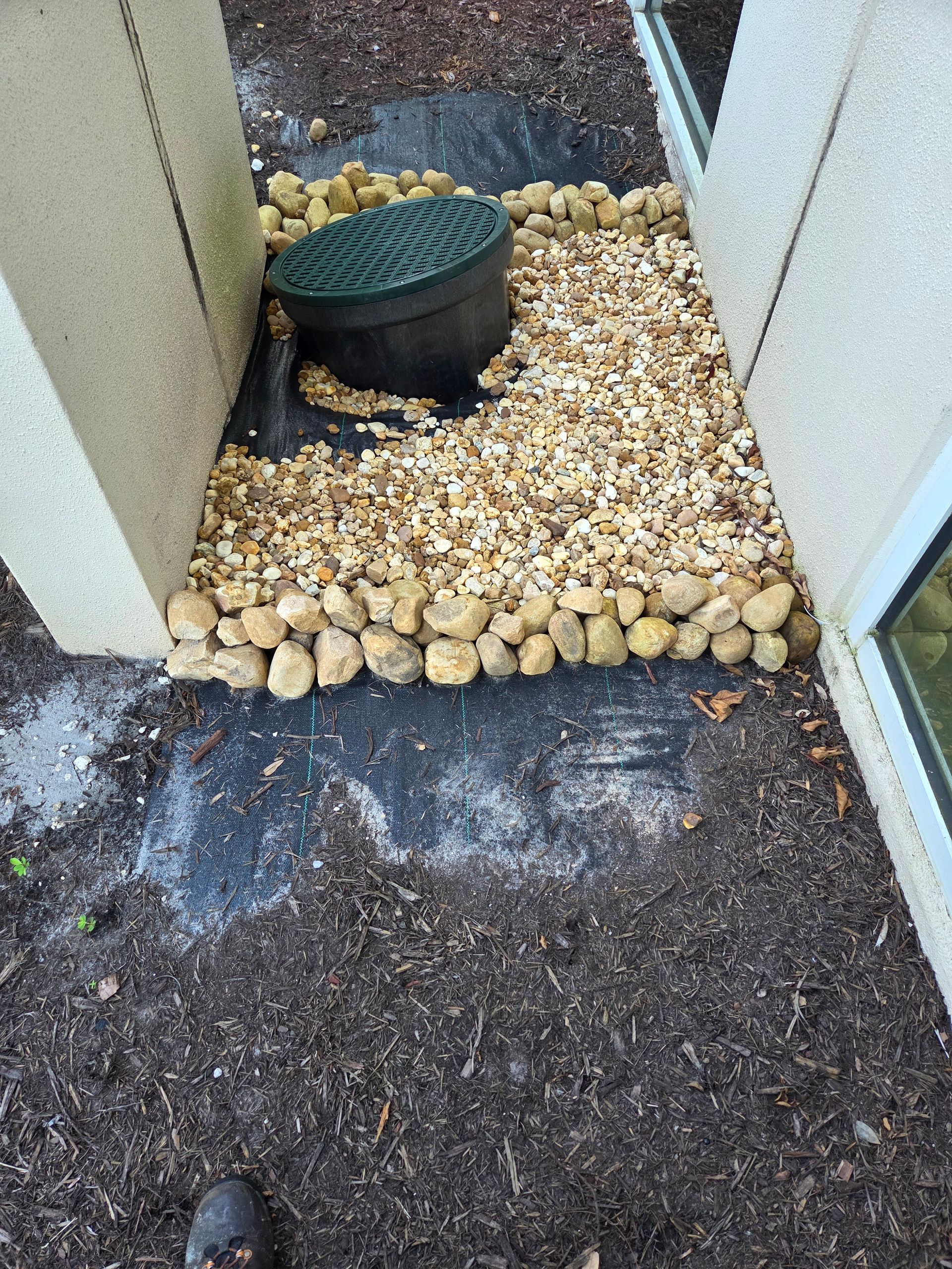 Gravel and rocks surrounding a drainage grate and between two walls. Black fabric is visible.