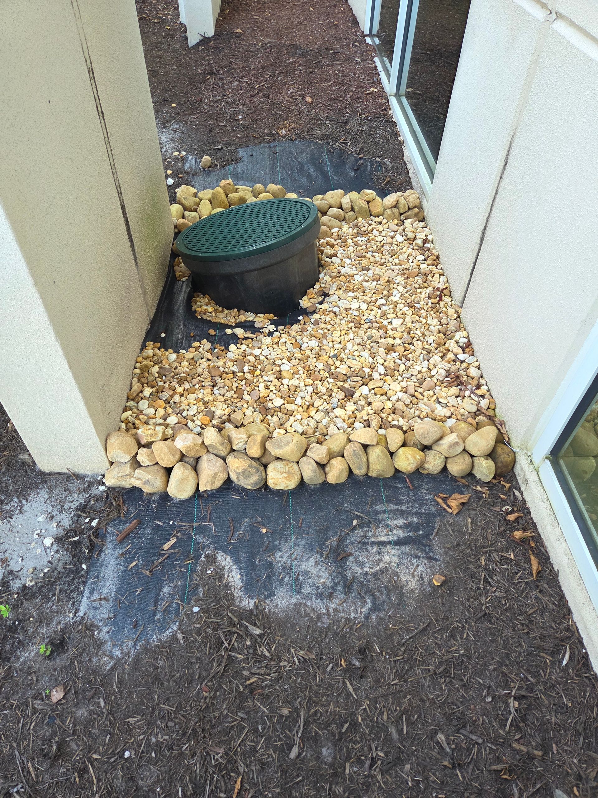 Gravel landscaping around a utility access point next to a building. Black weed barrier visible.