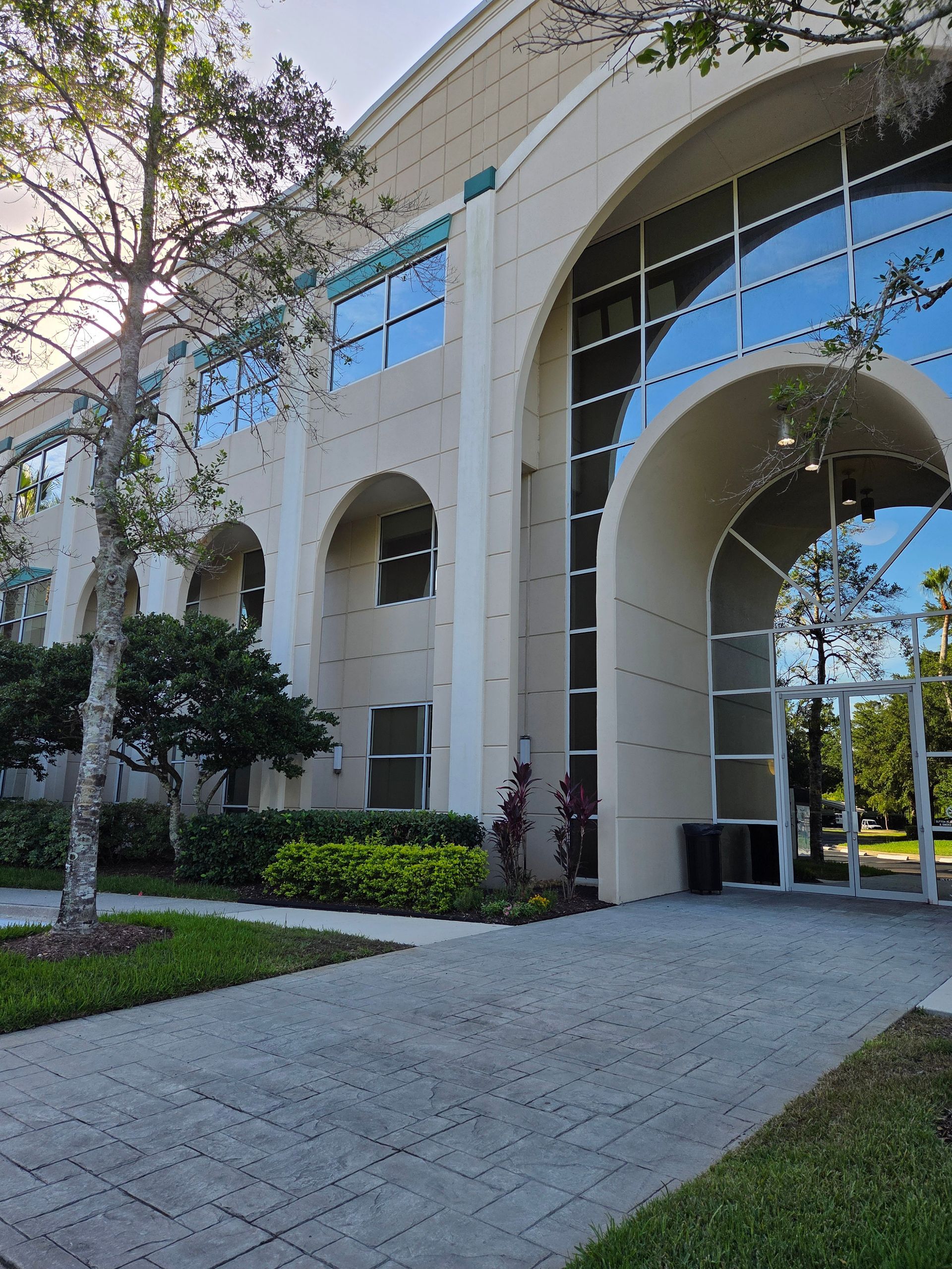 Beige building with arched windows and a glass entrance, sidewalk in the foreground, trees and bushes surrounding.
