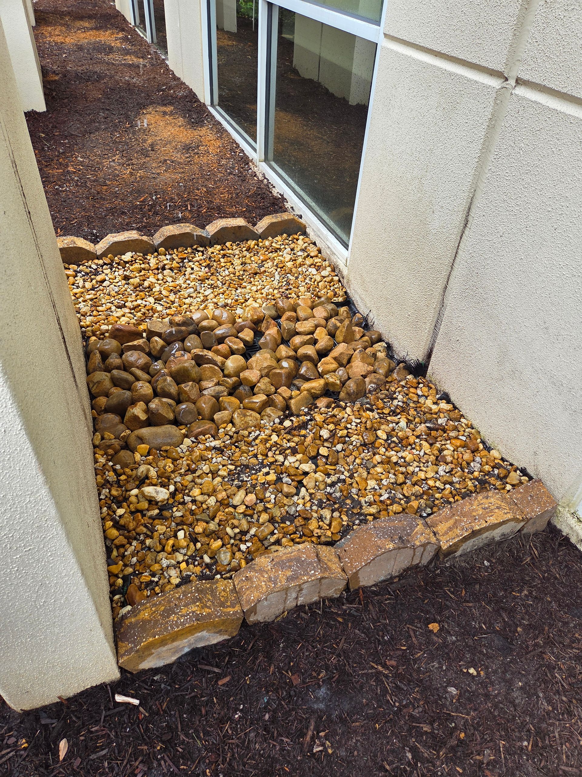 Landscaped area with brown mulch, pebbles, and rock edging next to a building's window.