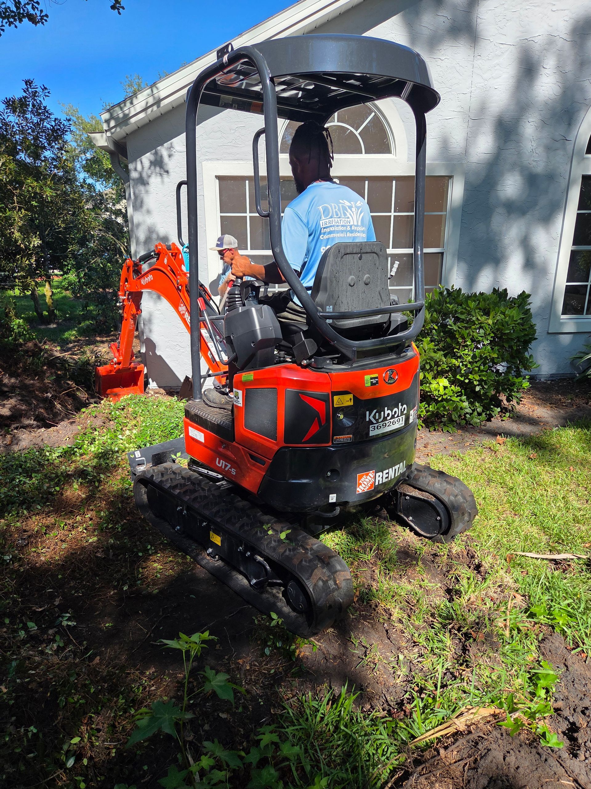 A small, red excavator operated by a person in a blue shirt is digging near a building.