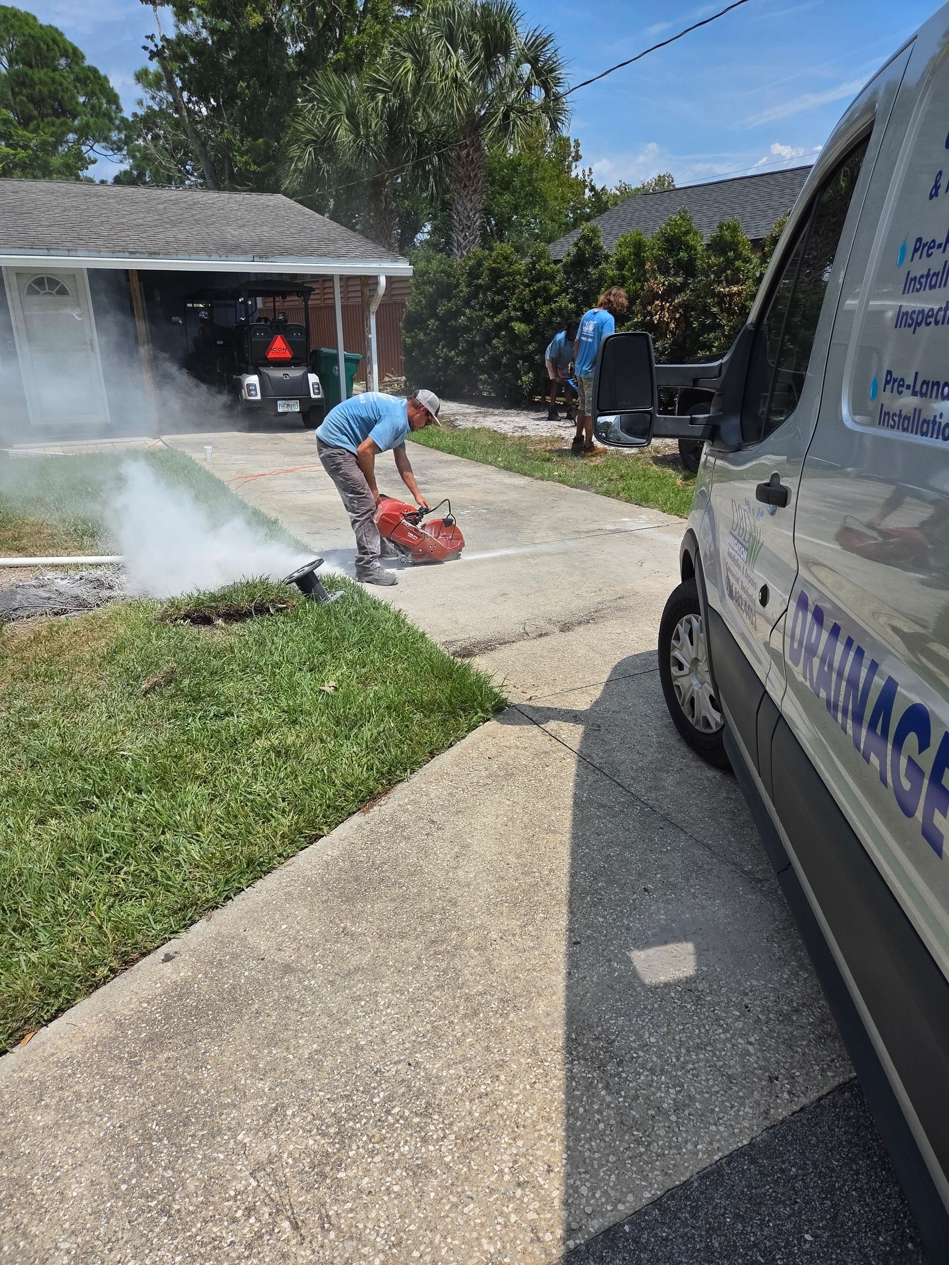 Man cutting concrete driveway, creating dust. Van with company logo parked nearby, workers in background. Sunny day.