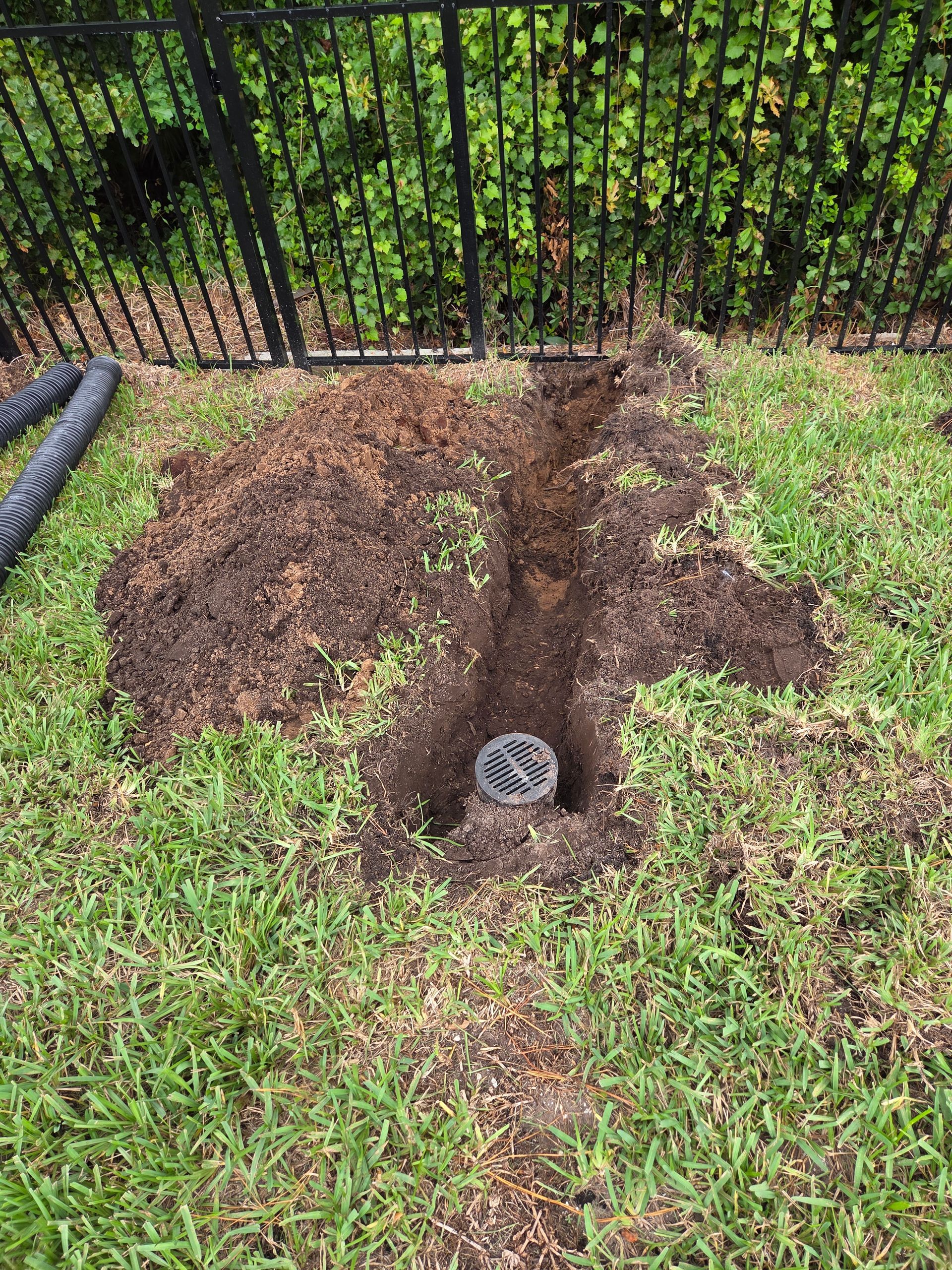 Hole dug in grass for a drain, with loose soil and a black fence in the background.