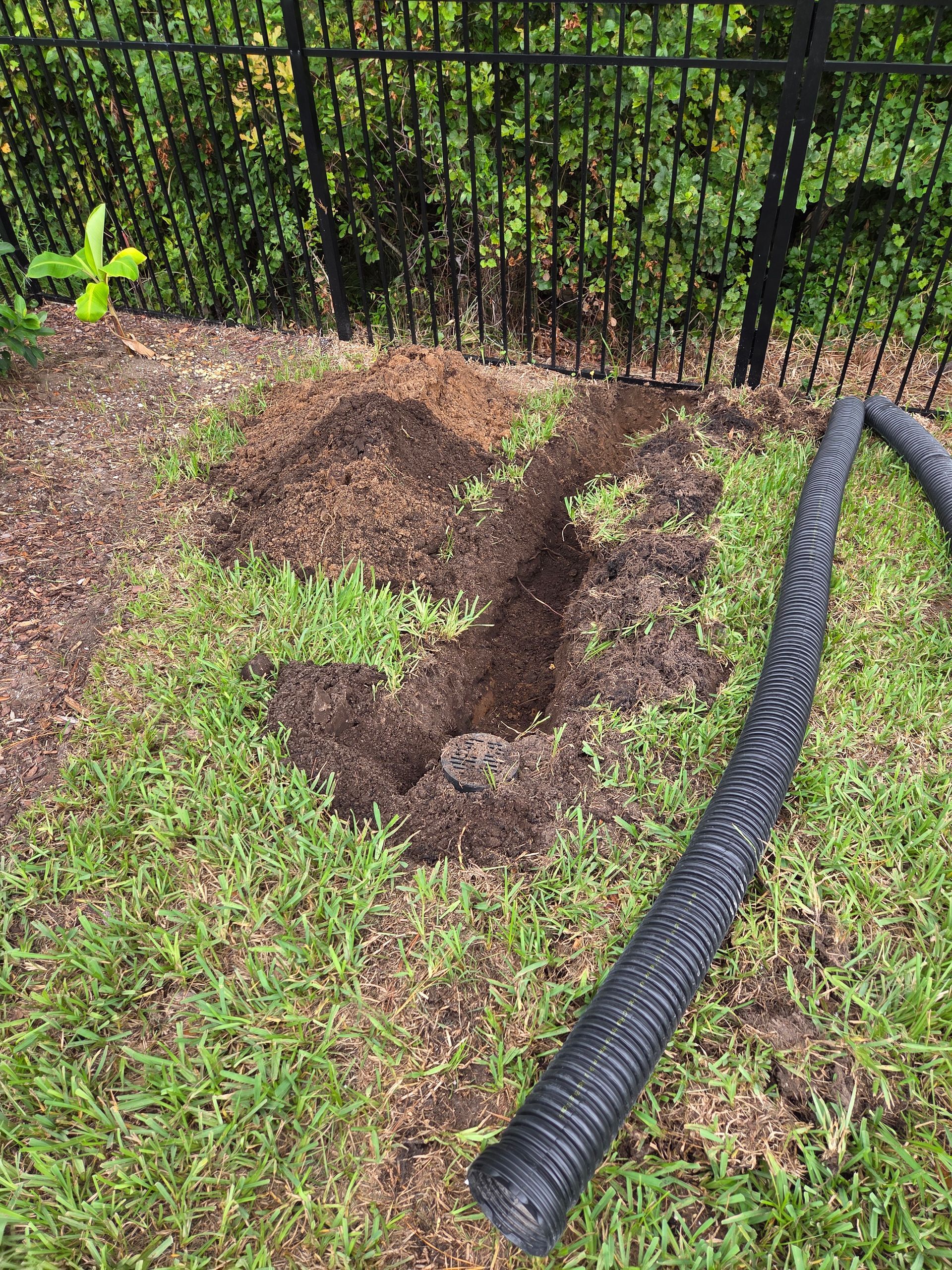 Trench dug in grass, beside black corrugated pipe, near a fence and pile of dirt.