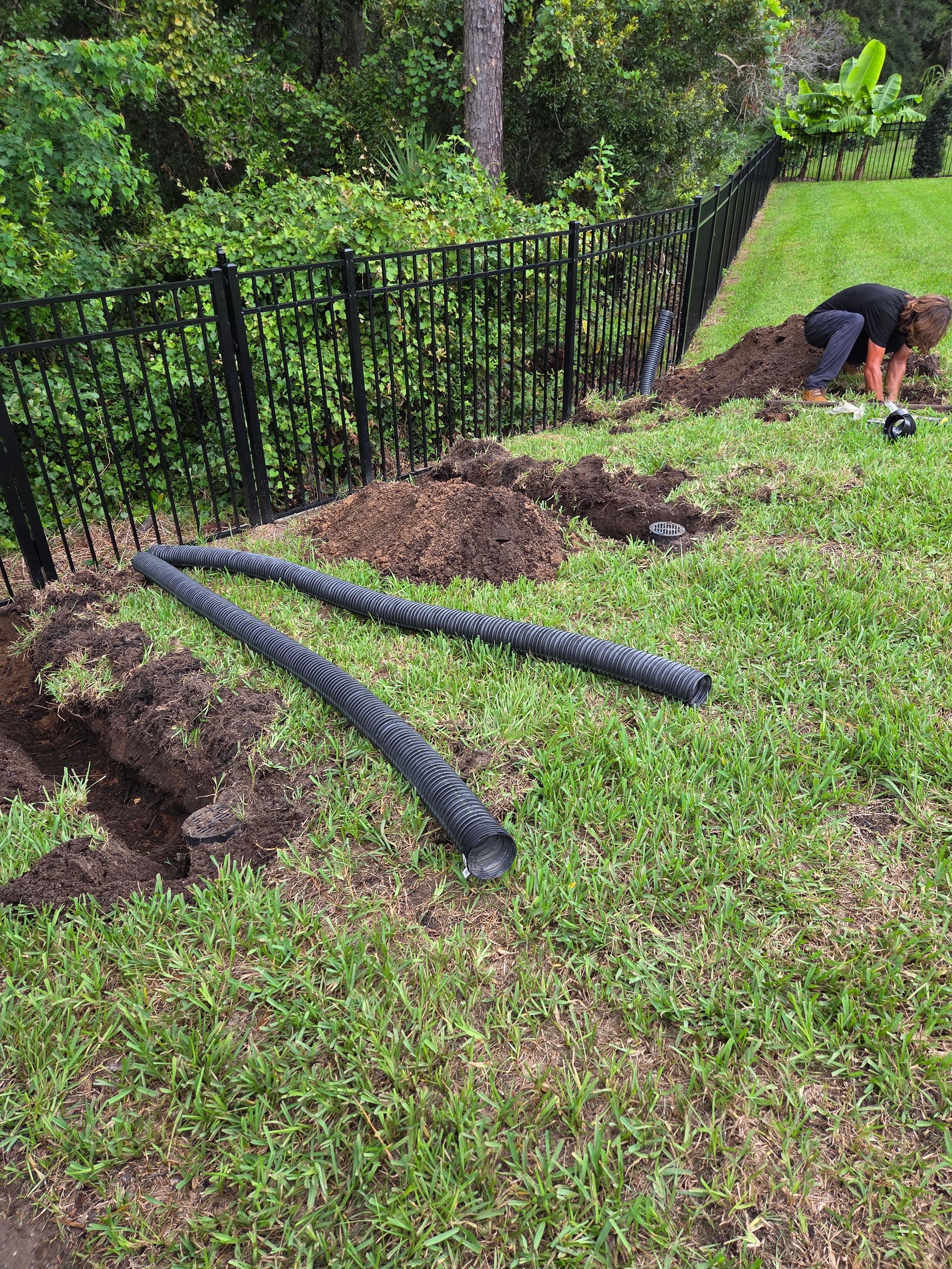 Black corrugated pipes and trench next to a fence with a person digging in the background. Green grass and foliage.