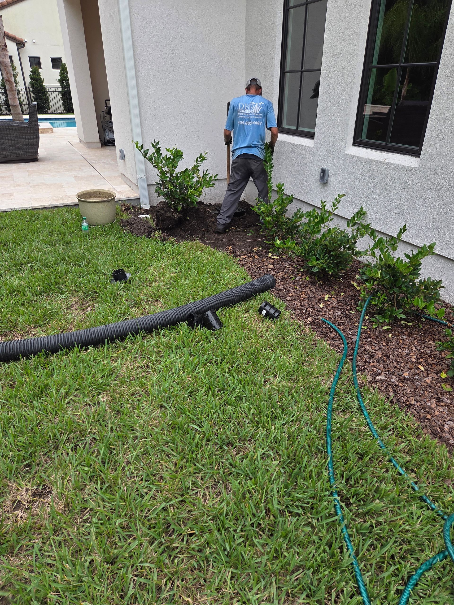 A person works on landscaping near a house, with green grass, bushes, and a drainage pipe visible.