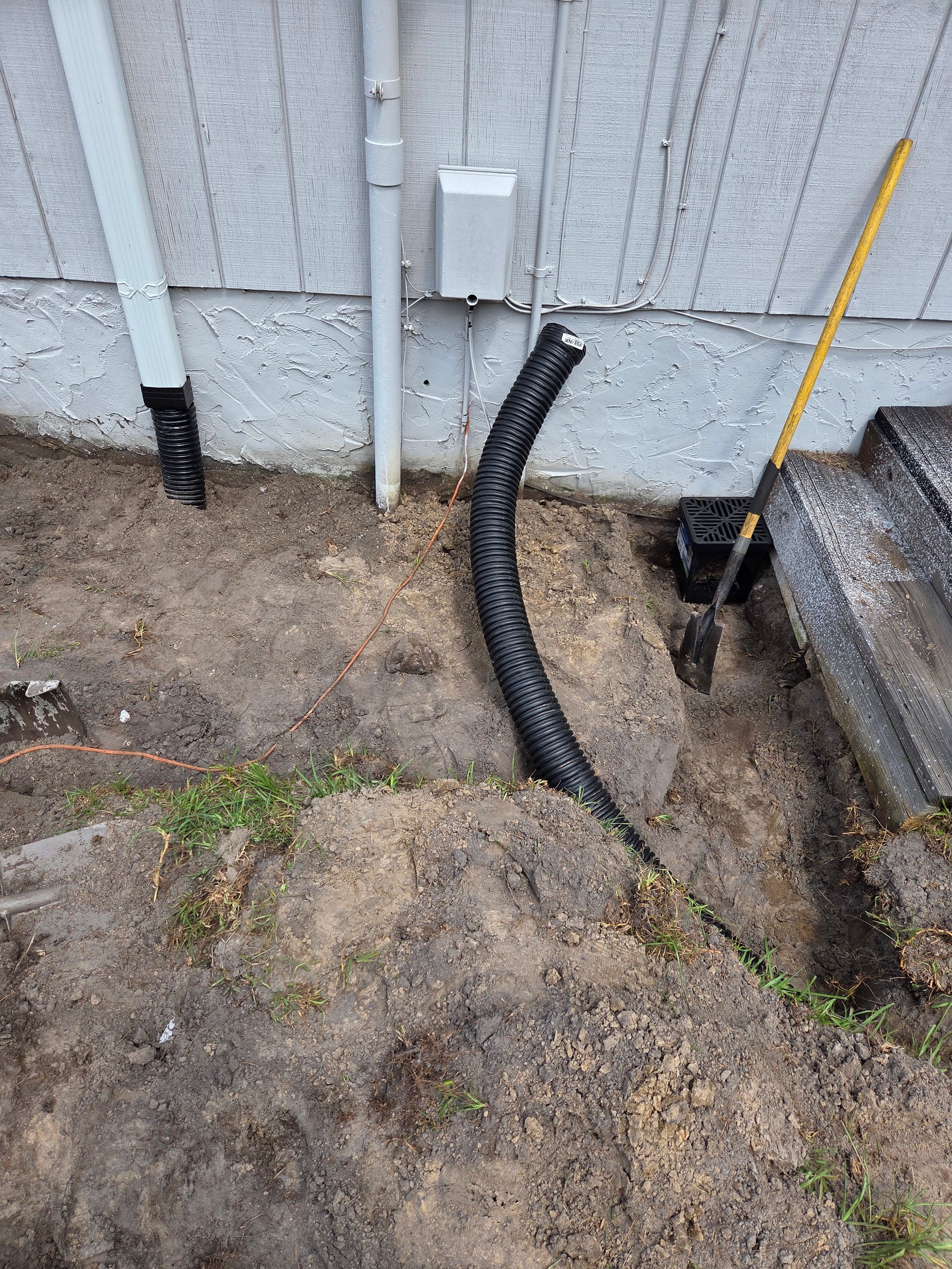 Black flexible conduit emerging from ground near a wall with electrical outlet. Spade and small wooden plank visible.