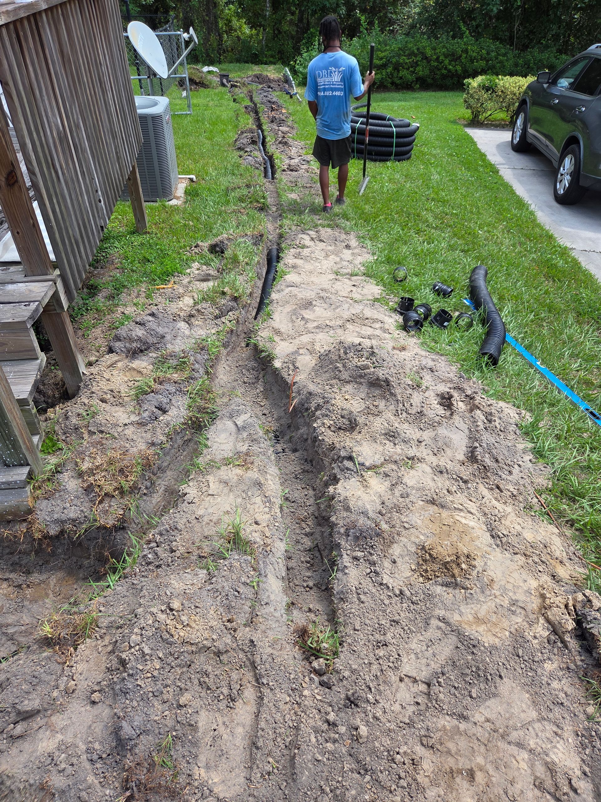 Man carrying black tubing walks next to a trench in a grassy yard.