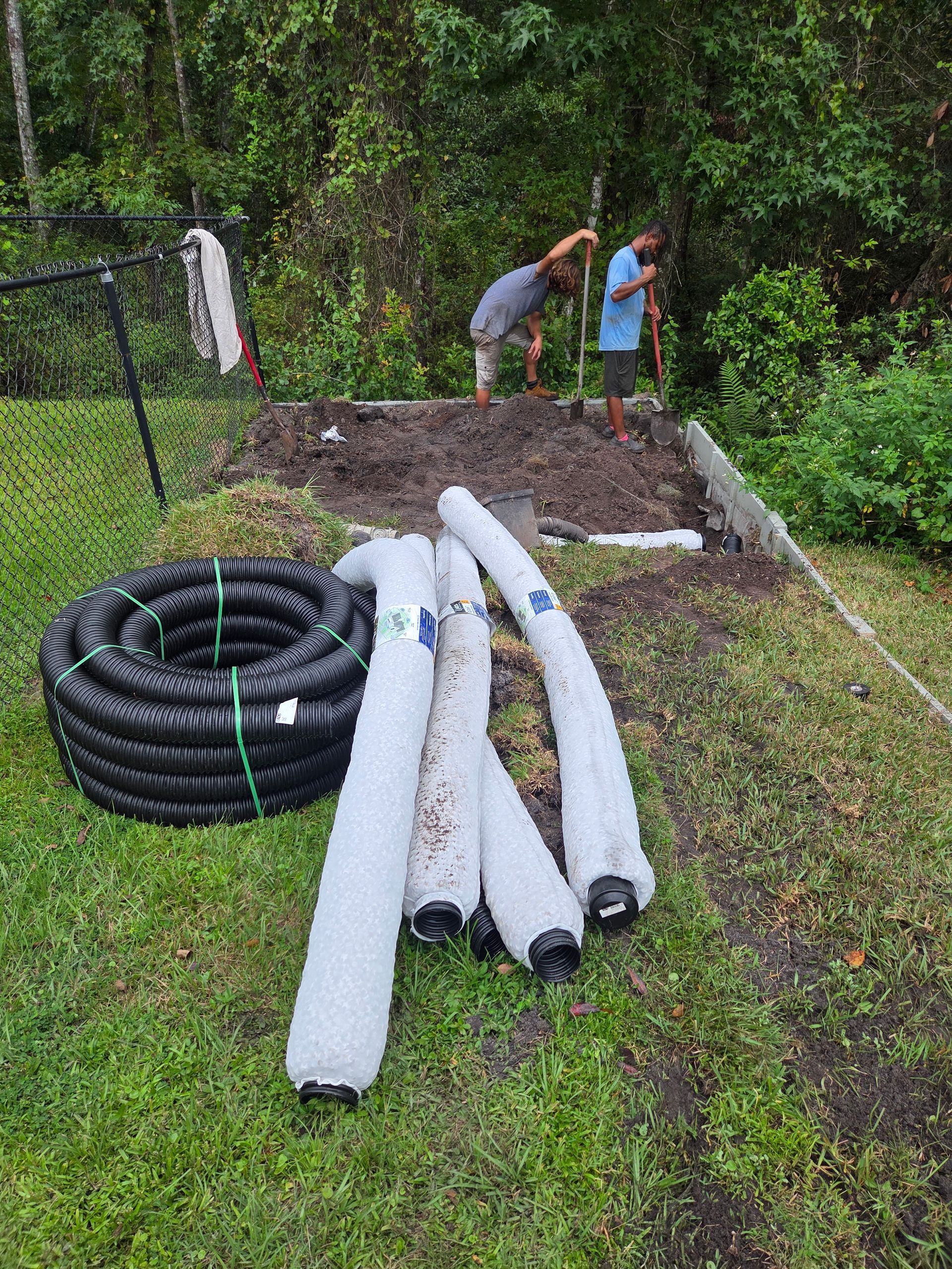 Two people digging in a garden bed, with coiled black drainage pipe and filter socks on the grass.