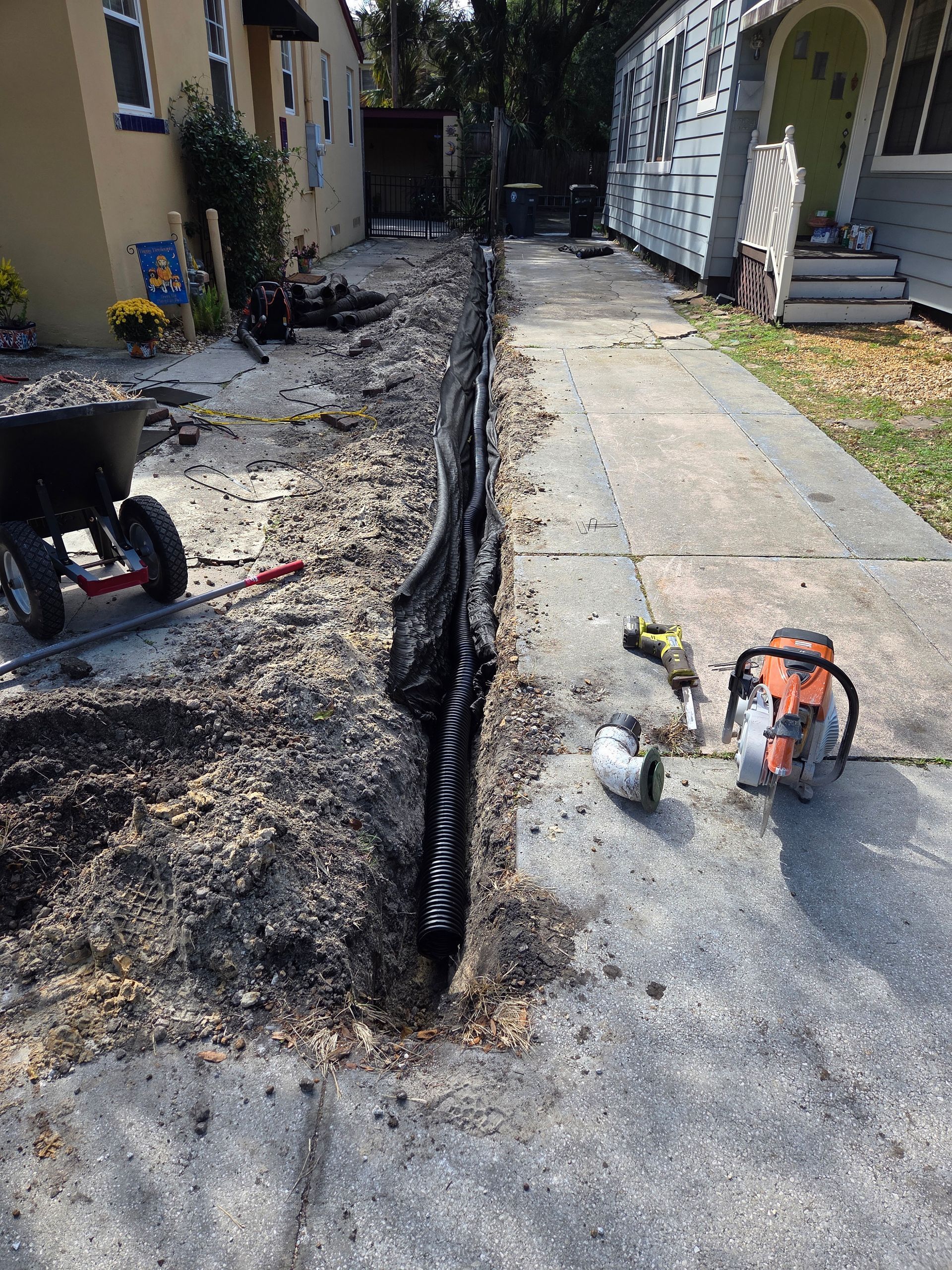 Trench dug in a driveway with pipe and tools for installation. Buildings in background.