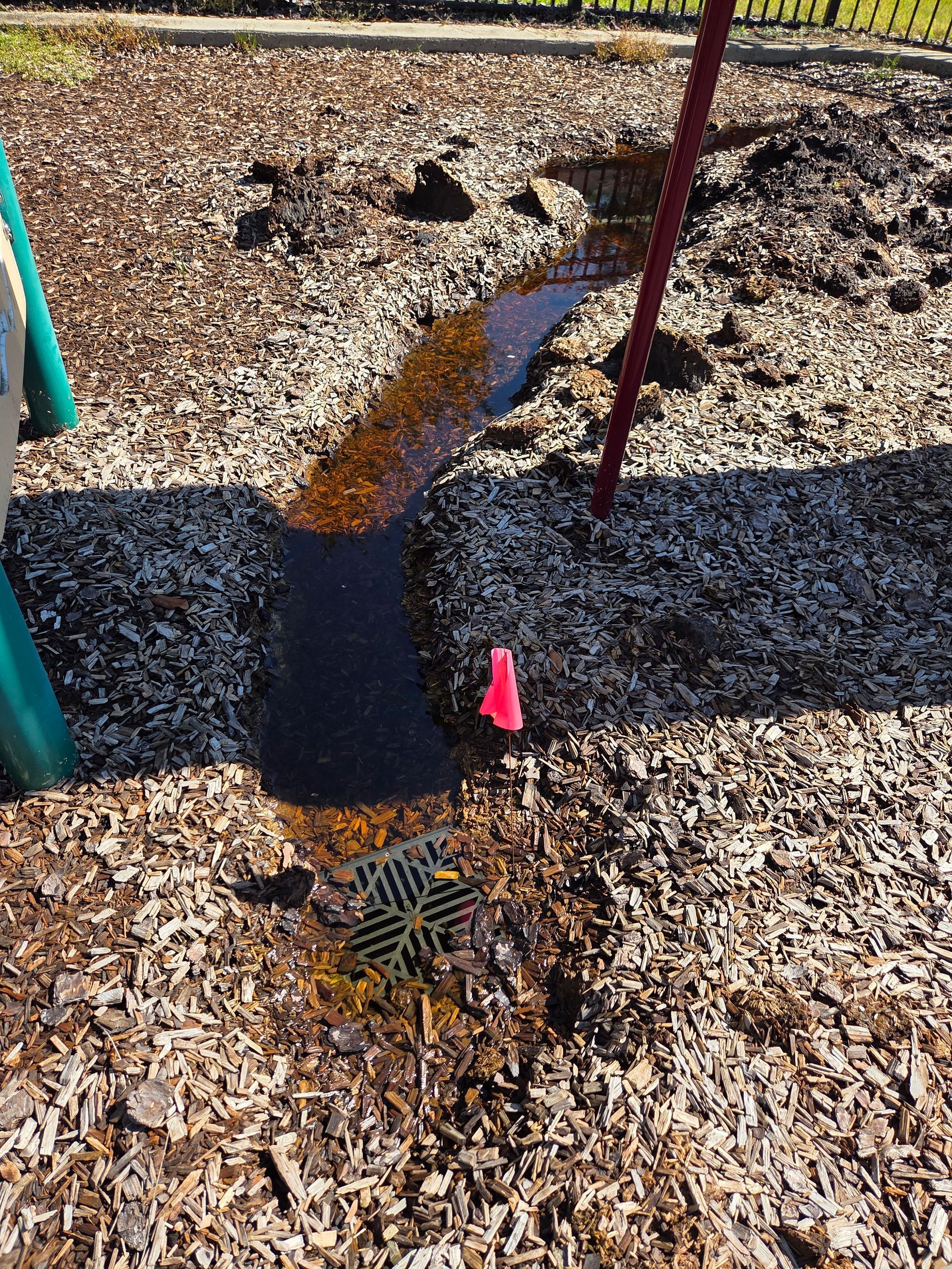 Water flows across wood chips toward a drain at a playground. A pink flag is next to the drain.