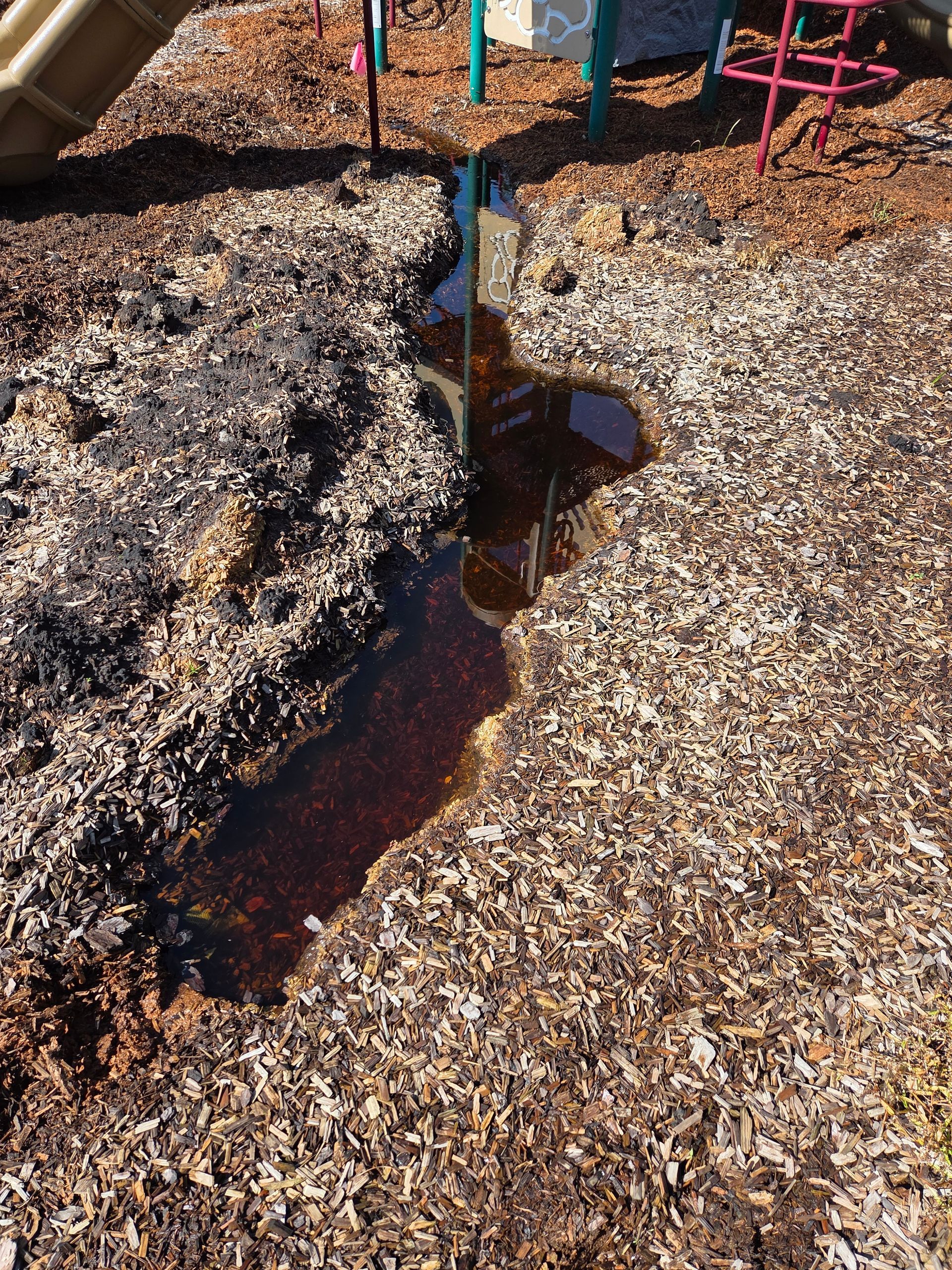 Puddle of dark liquid on wood chips at a playground. Water runs along a trench-like path.