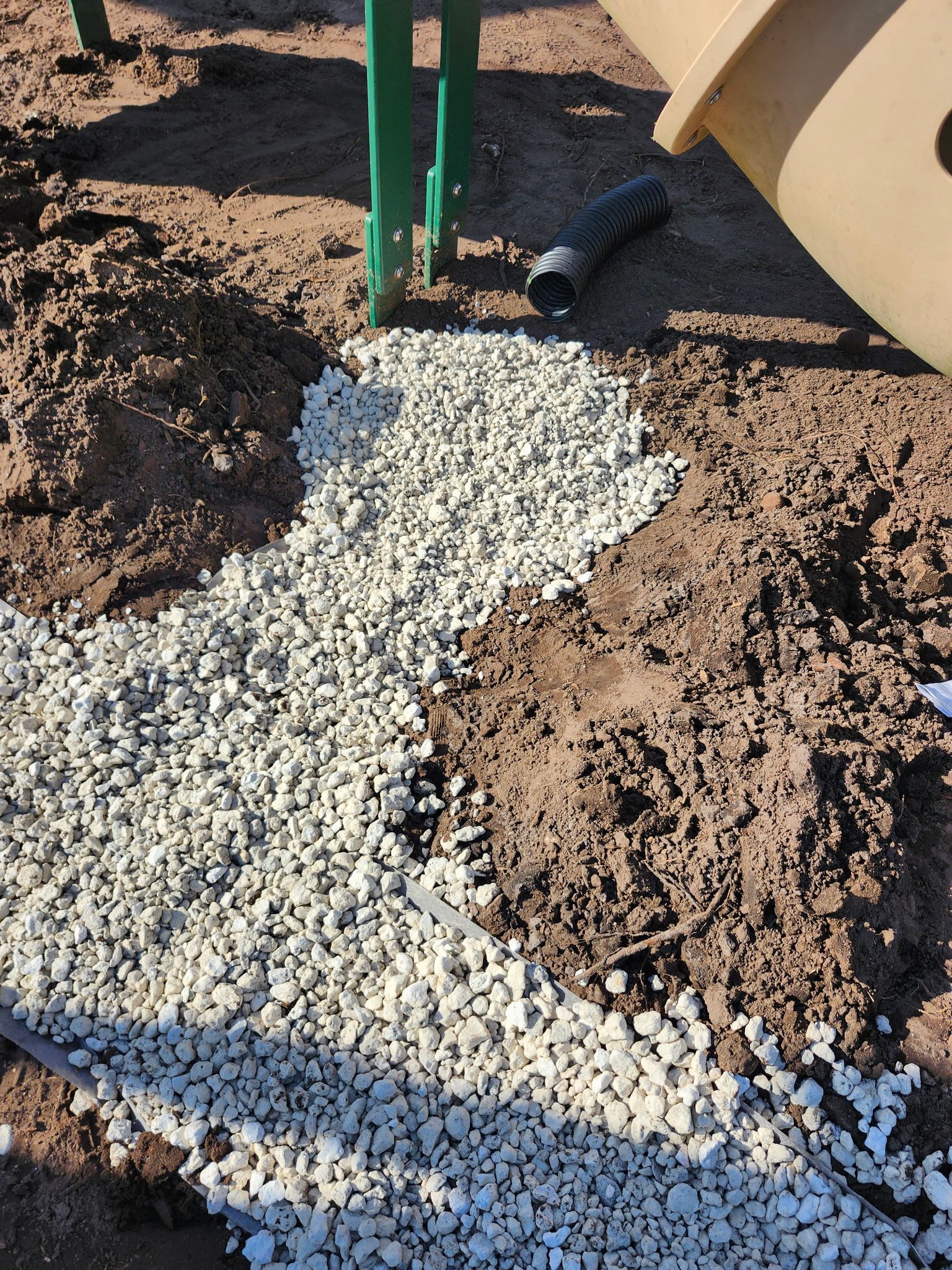 Gravel pathway under playground equipment, contrasted with dirt and a black drainage pipe.