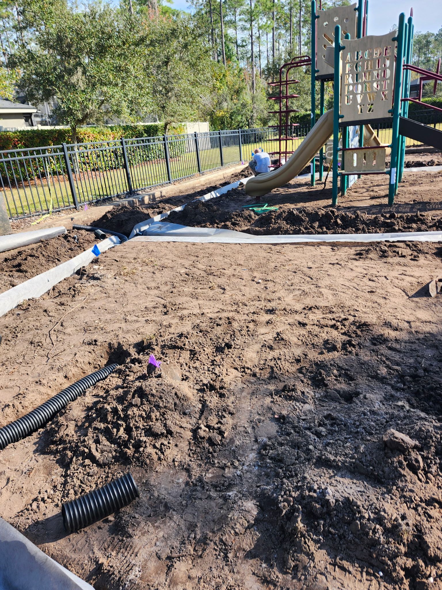 Playground construction in progress; exposed soil, drain pipes, and partially built play structure.