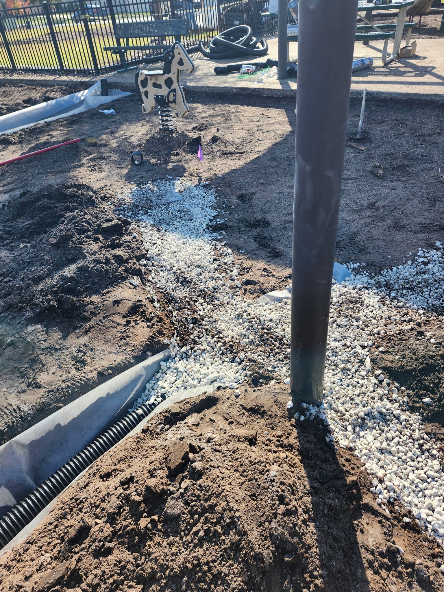 Construction site with dark pole, gravel, soil, and drainage piping. Playground equipment visible in background.