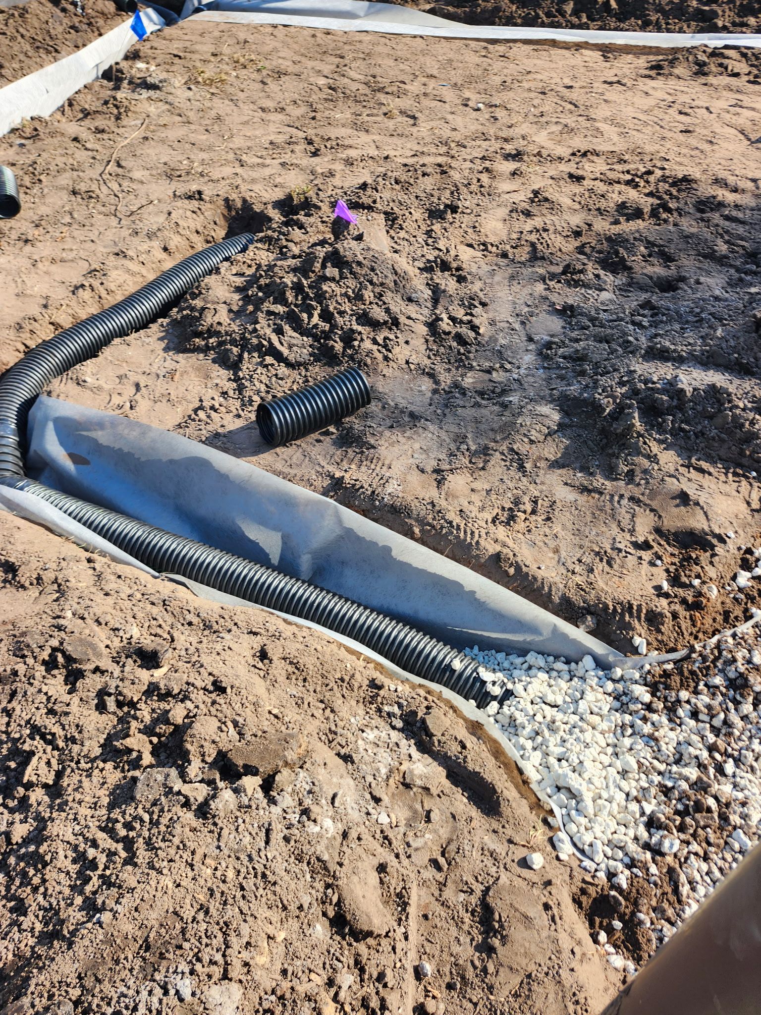 Trench filled with gravel, wrapped in fabric, with black drainage pipe, in a construction site.