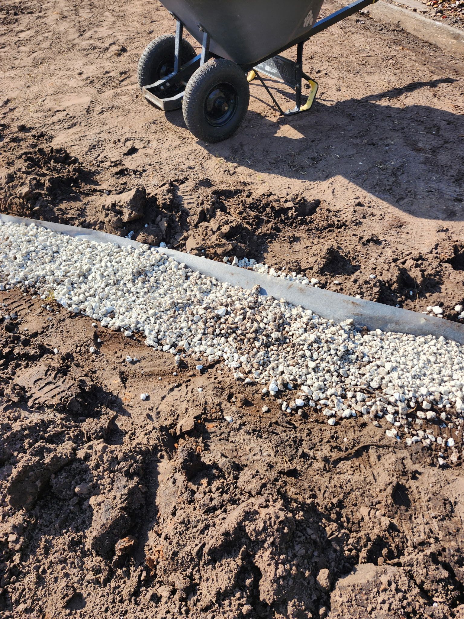 Wheelbarrow next to a gravel-filled trench. Dark soil surrounds the gravel, outdoors.