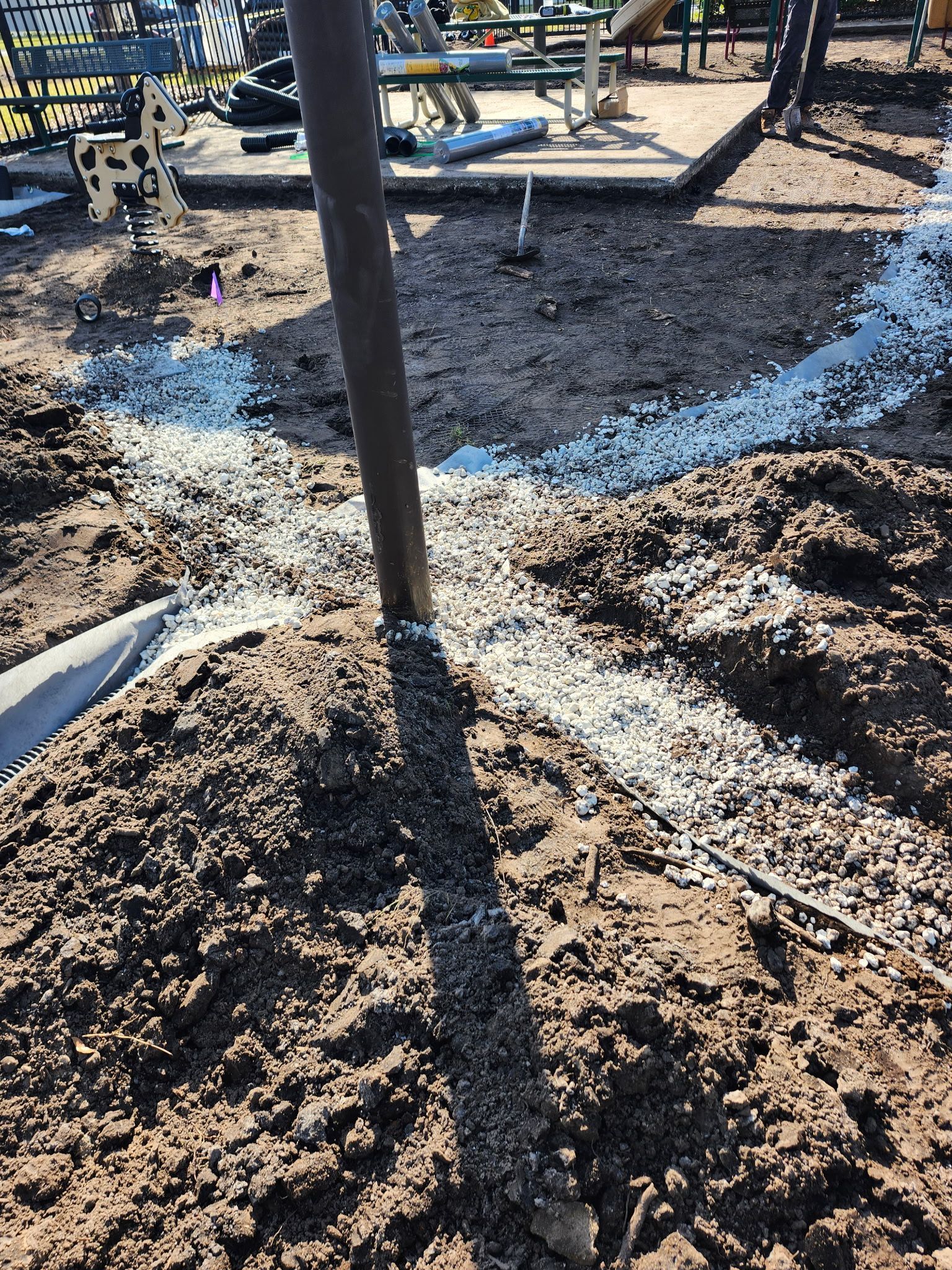 A playground under construction. A dark pole is centered, with gravel lines radiating outward in the dirt.