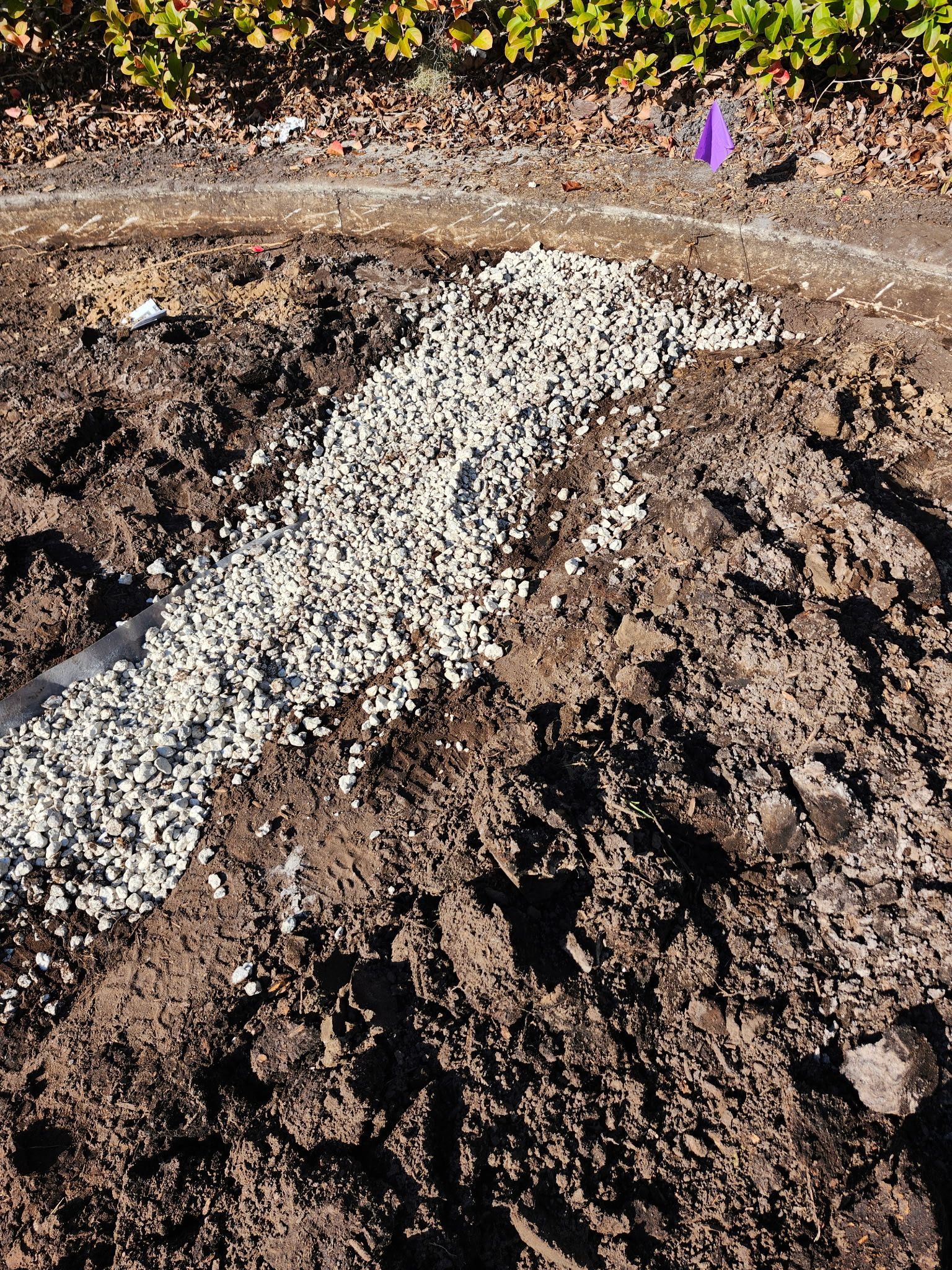 Gravel-filled trench in dirt, purple marker in background, top edge has plant growth.