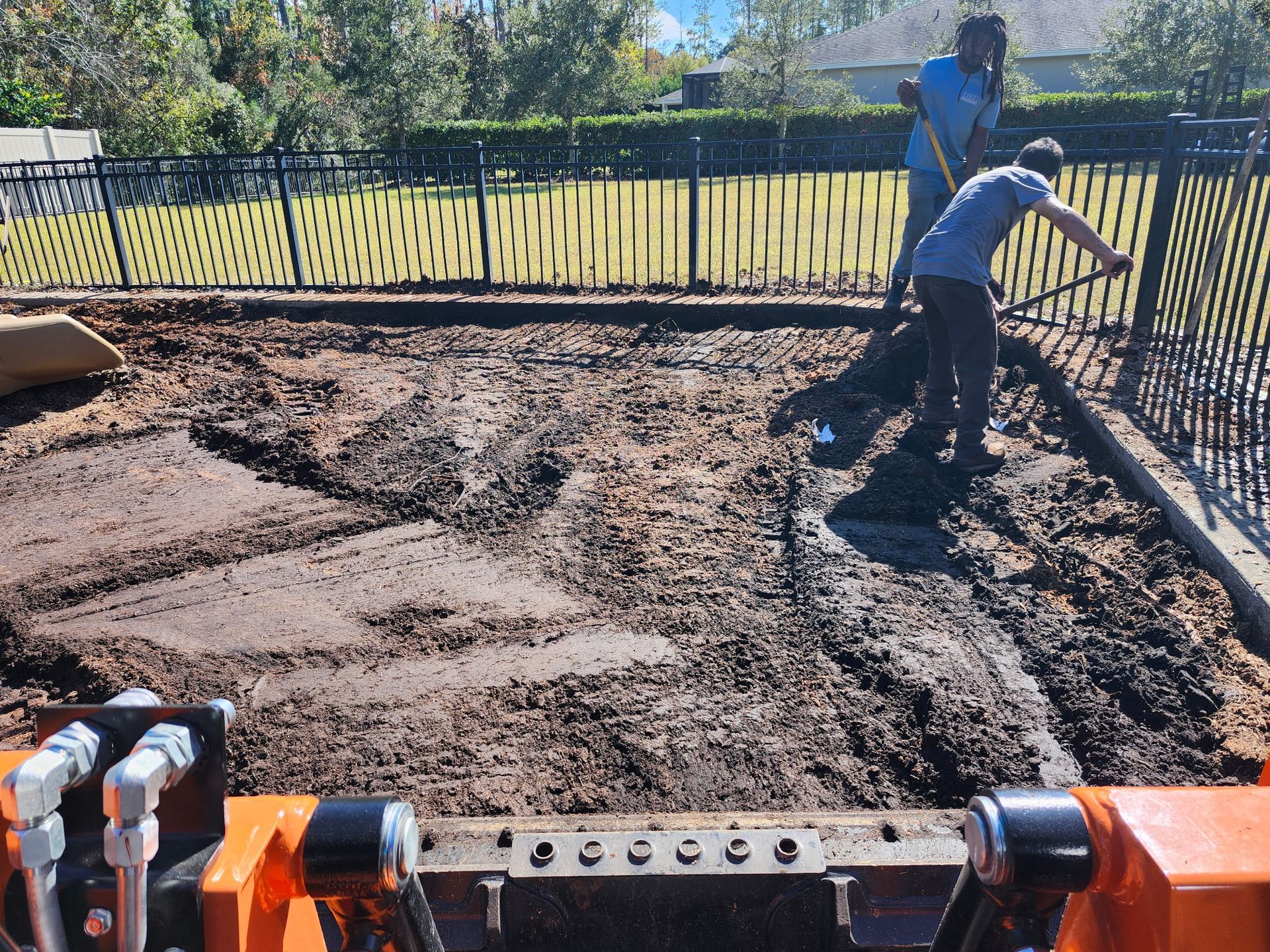 Two people leveling soil in a dirt plot, viewed from a tractor bucket. Fence and trees in background.