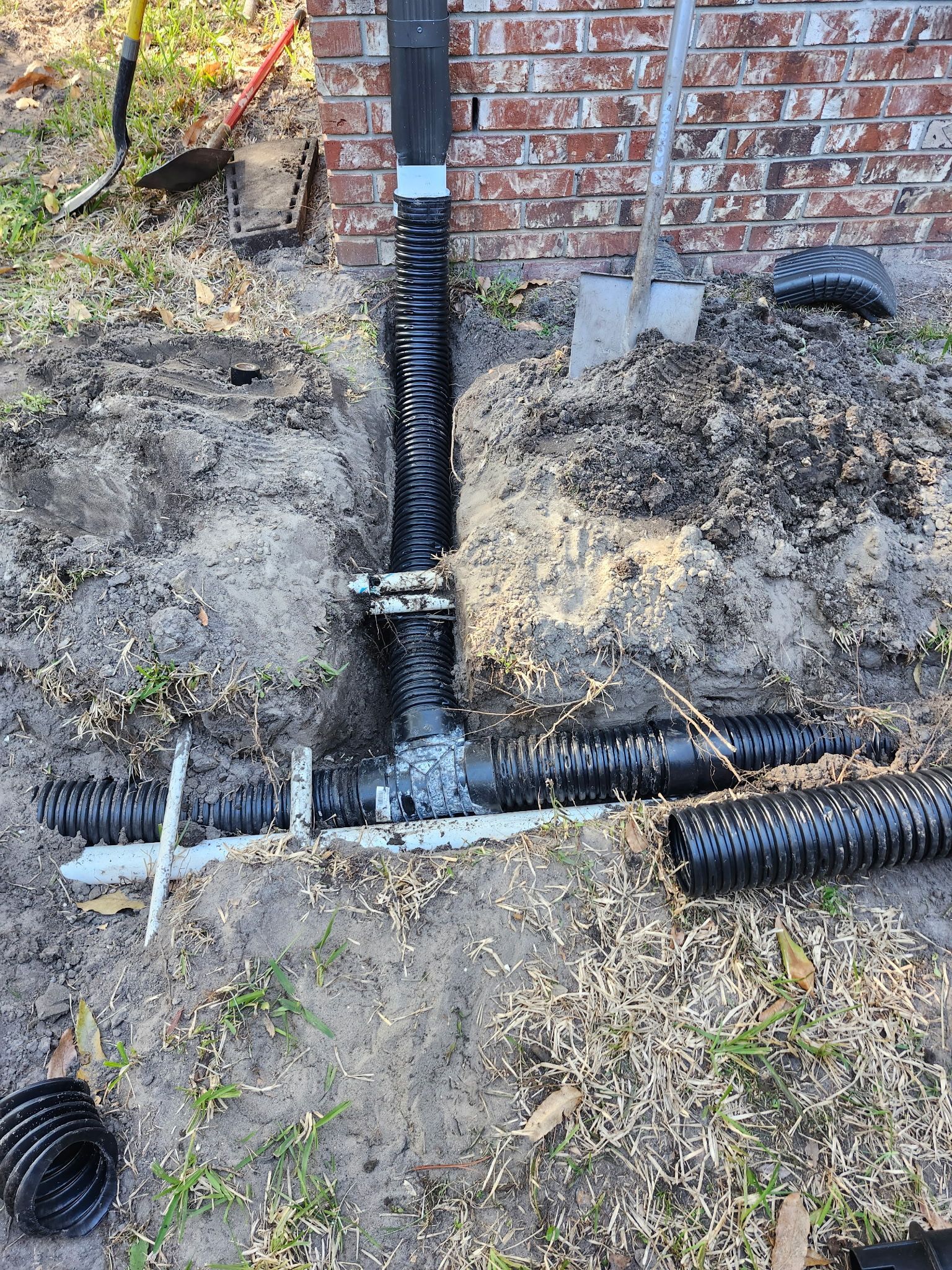 Black corrugated drain pipes installed near a brick wall, dirt, shovel, and grass are present.