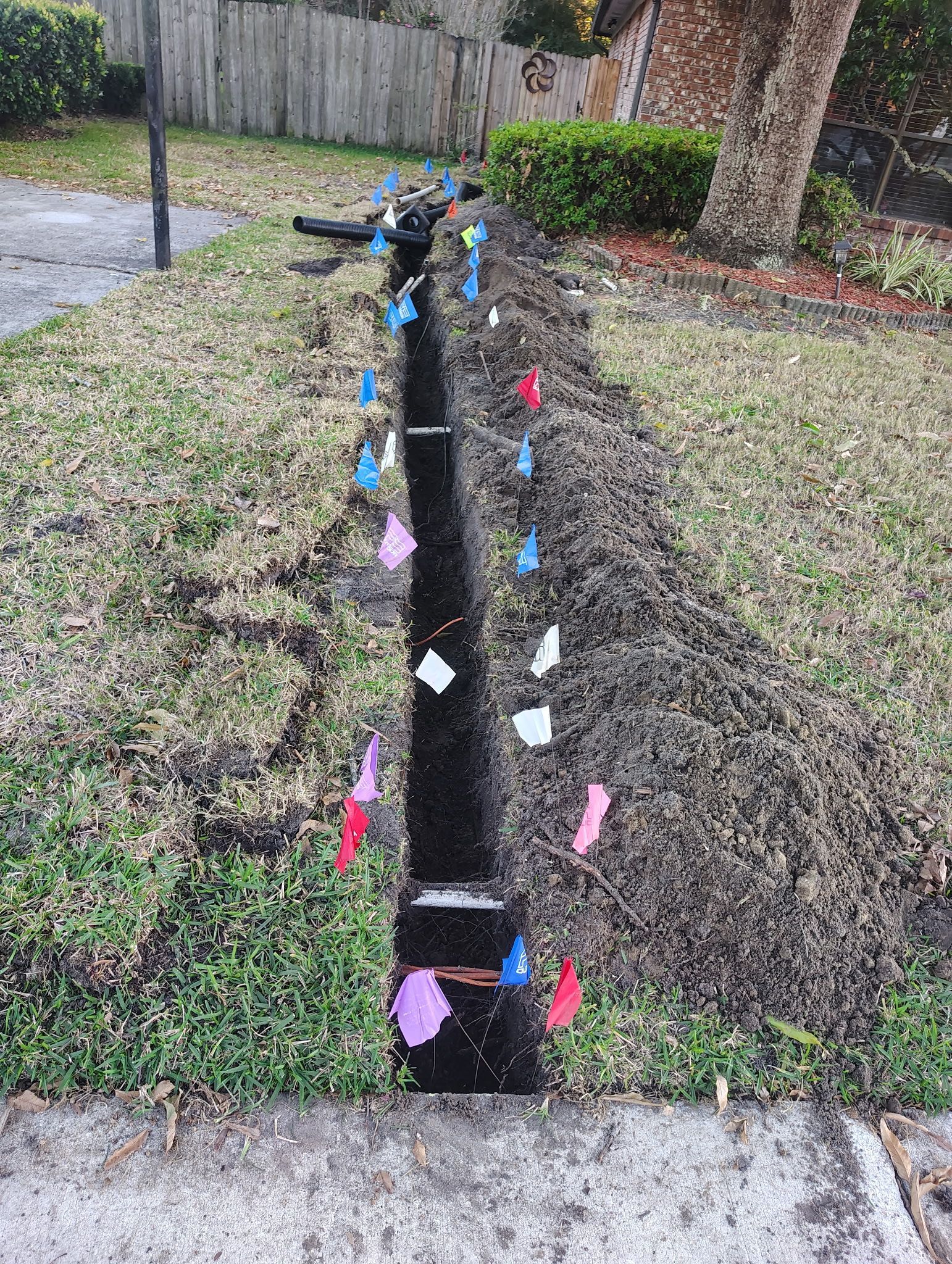 Trench in yard with colored flags marking buried utility lines, bordered by grass and sidewalk.