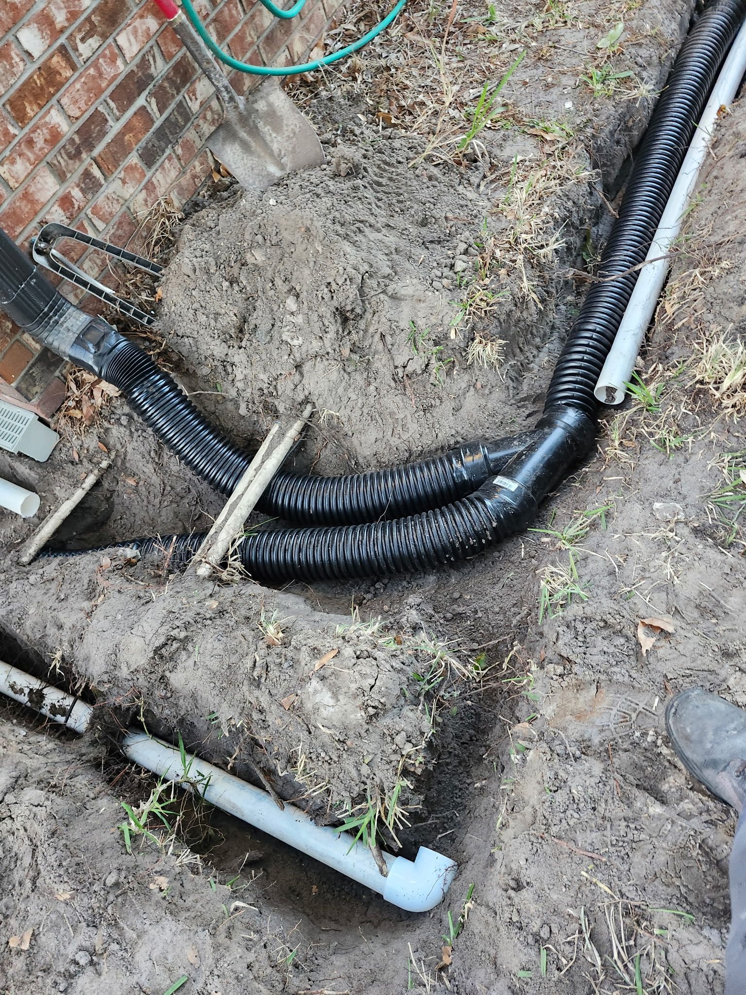 Pipes installed in a trench near a brick wall, likely for drainage. Black corrugated and white PVC pipes are visible.