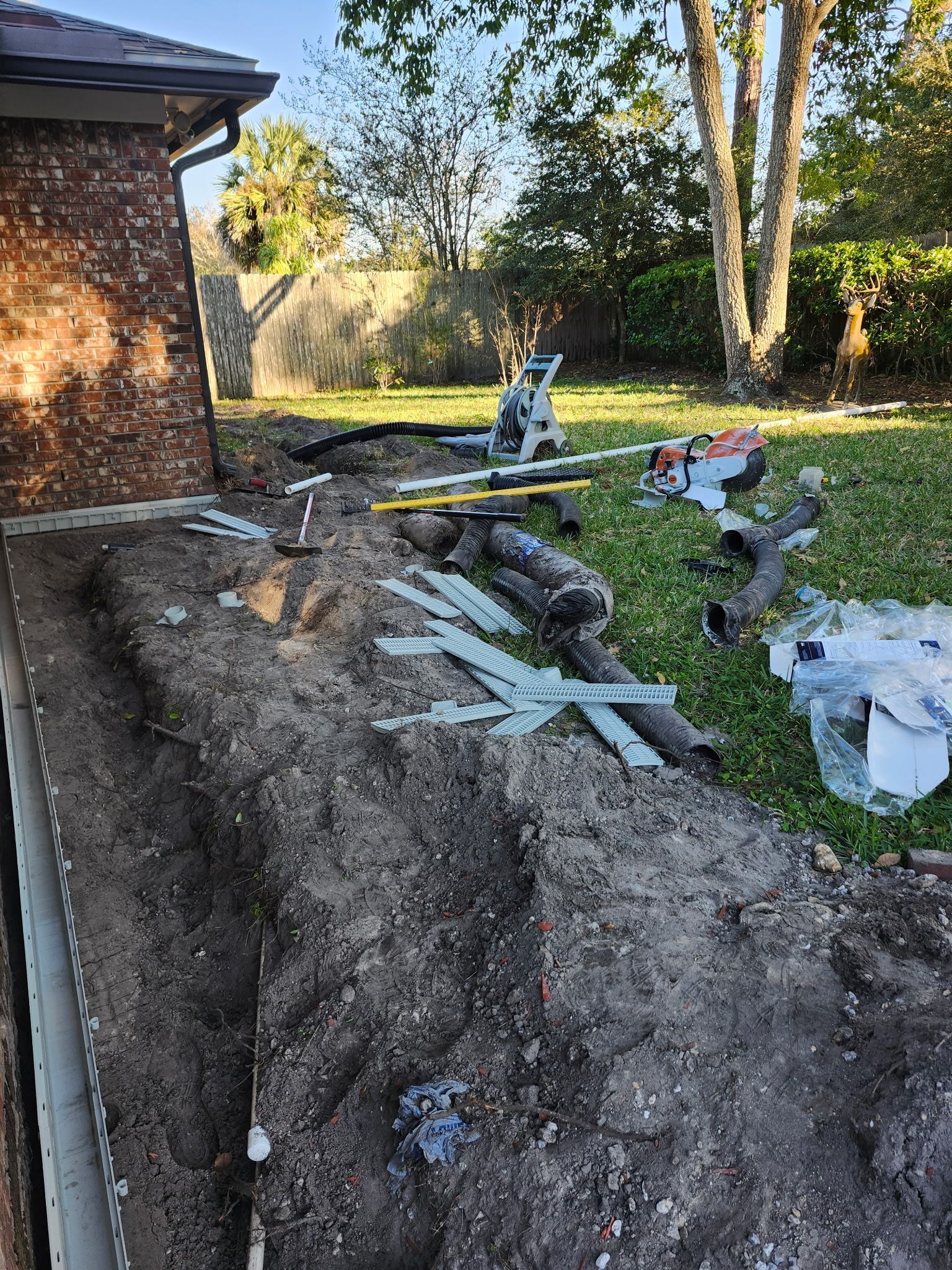 A trench along a brick building, with construction materials and saw on the grass.