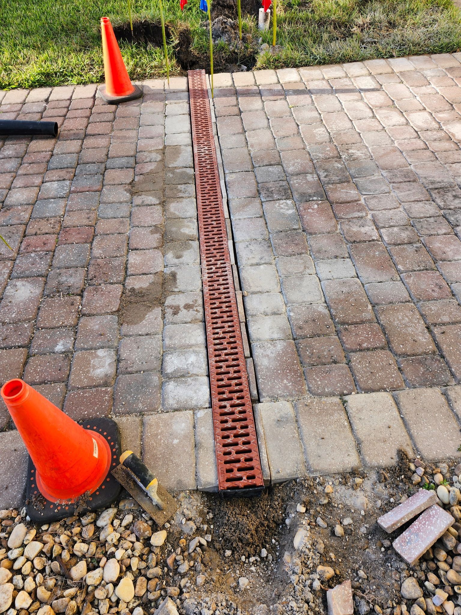 Brick paver driveway with trench and installed linear drain. Orange cones mark the construction zone.