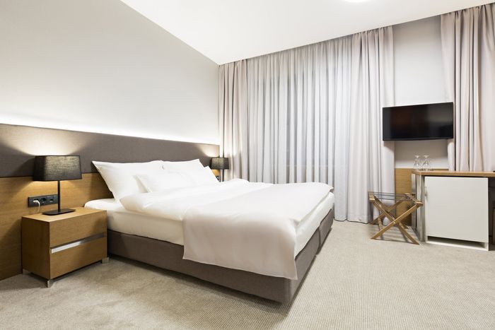 Neutral toned bedroom, with black lamp on timber bedside table. TV mounted on wall between set of neutral tone closed curtains.