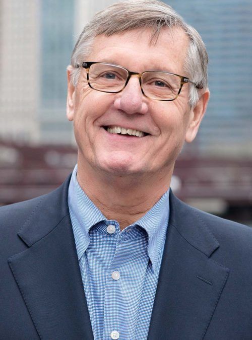 Smiling man in glasses and a blue shirt, wearing a dark blazer outdoors against a blurred city background